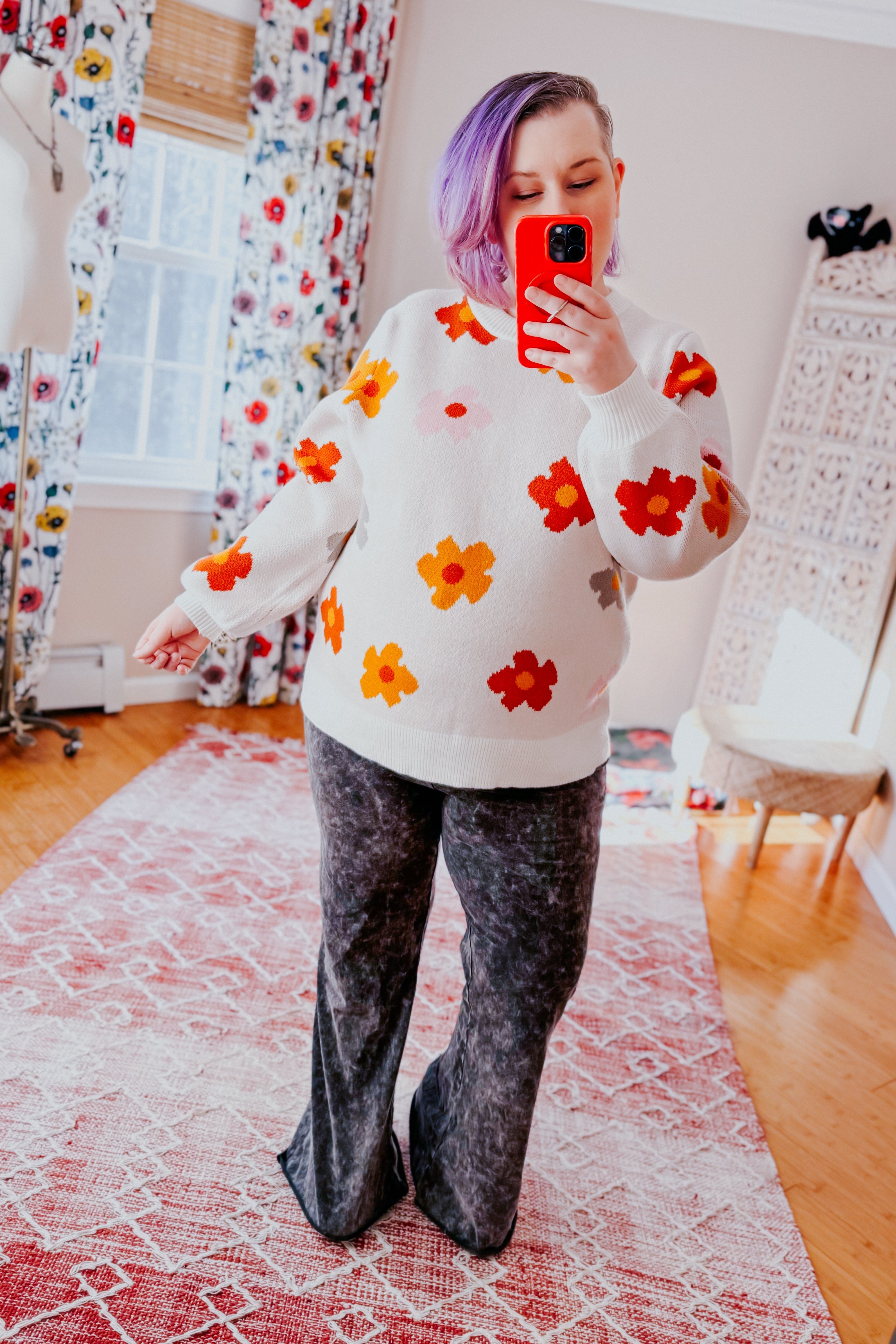 Woman with purple hair wearing white sweater with orange and red floral print and black wide-leg pants indoors