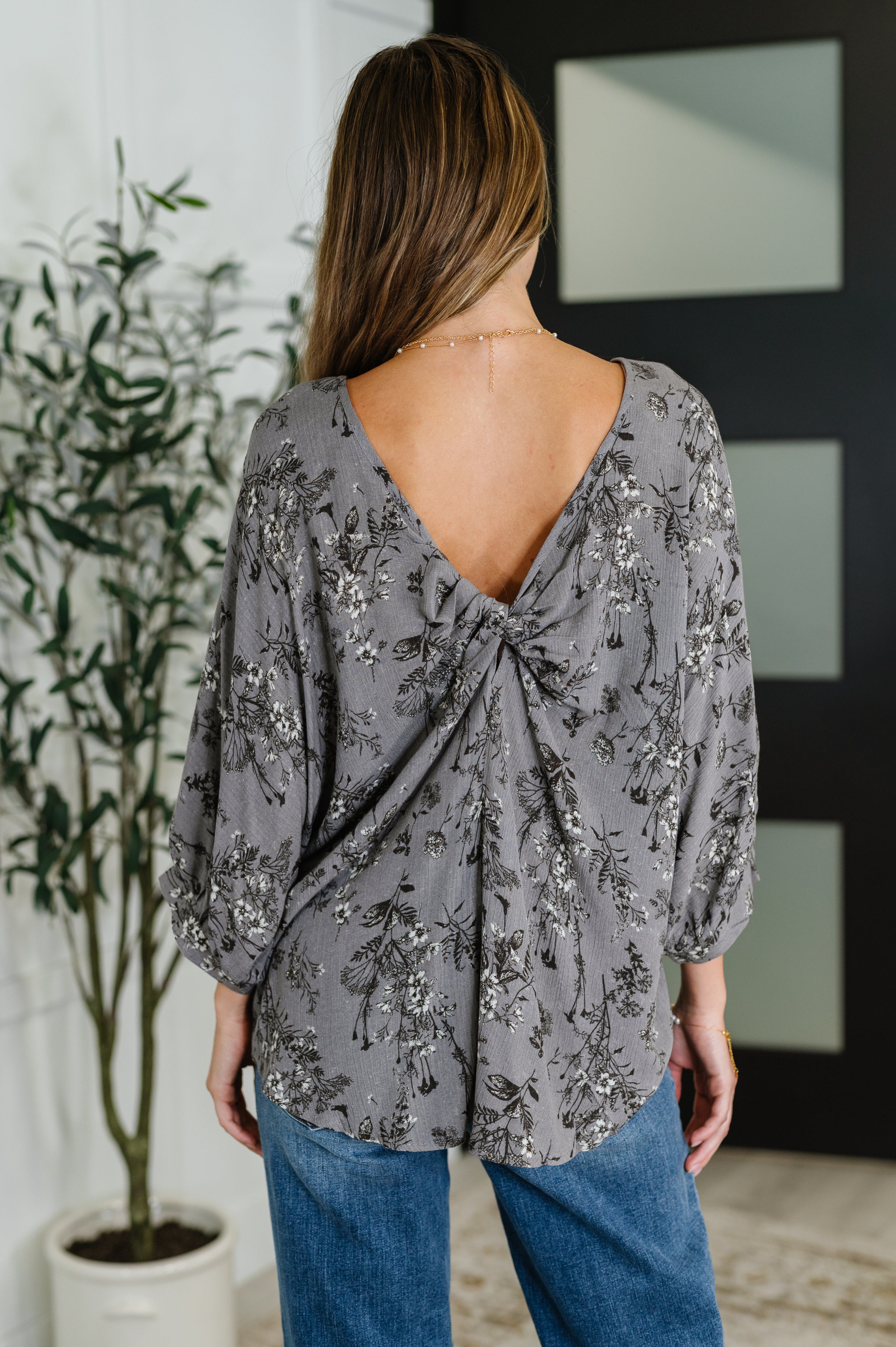 Woman with long brown hair wearing dune grey floral crinkle blouse with twisted deep V-back and blue jeans indoors