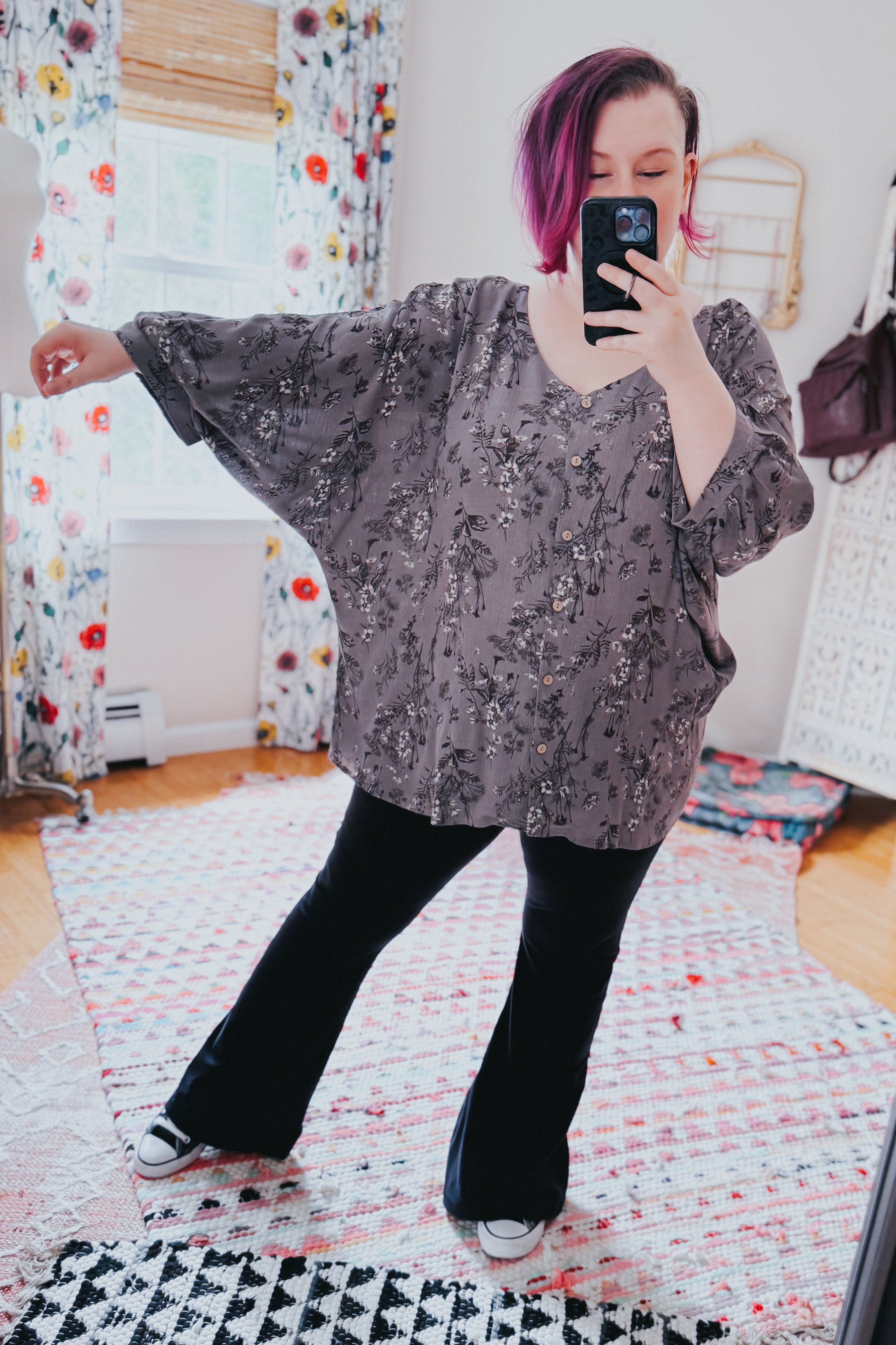 Woman wearing loose dune grey crinkle blouse with black floral print and black pants taking mirror selfie indoors
