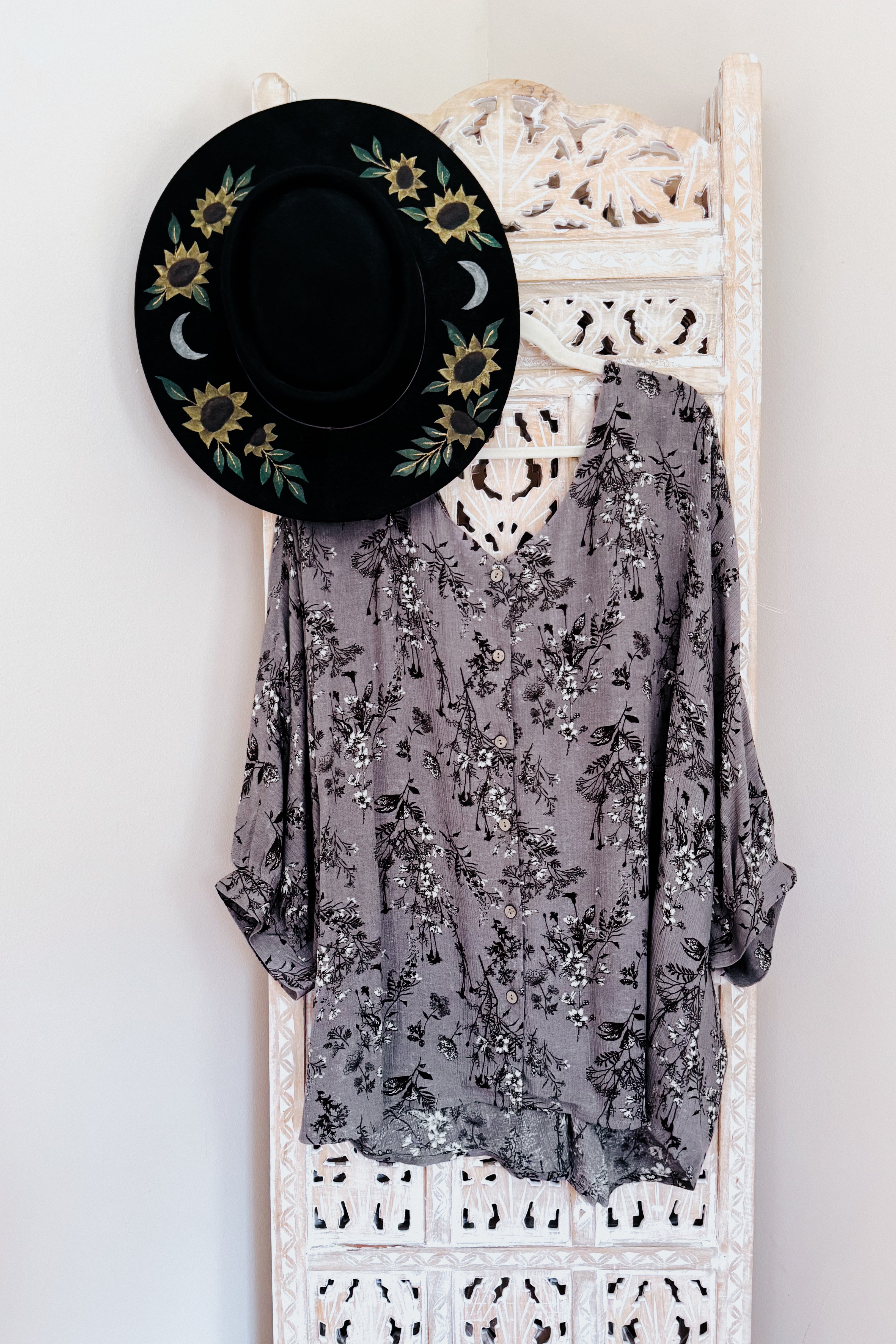 Dune grey crinkle woven blouse with black and white floral print hanging on carved white wooden screen with black hat