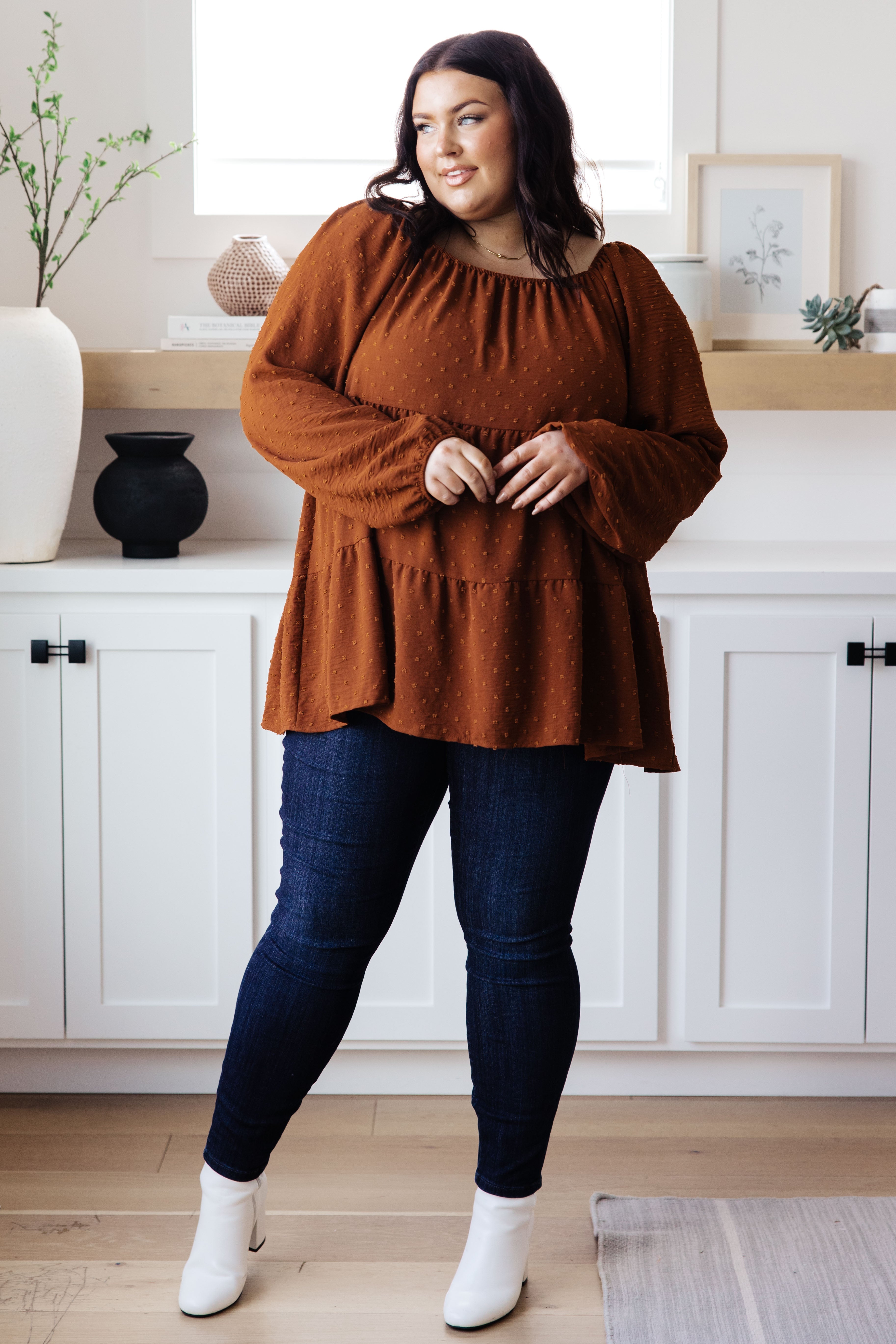 Plus-size model wearing rust tiered babydoll blouse with textured dot pattern and dark skinny jeans indoors