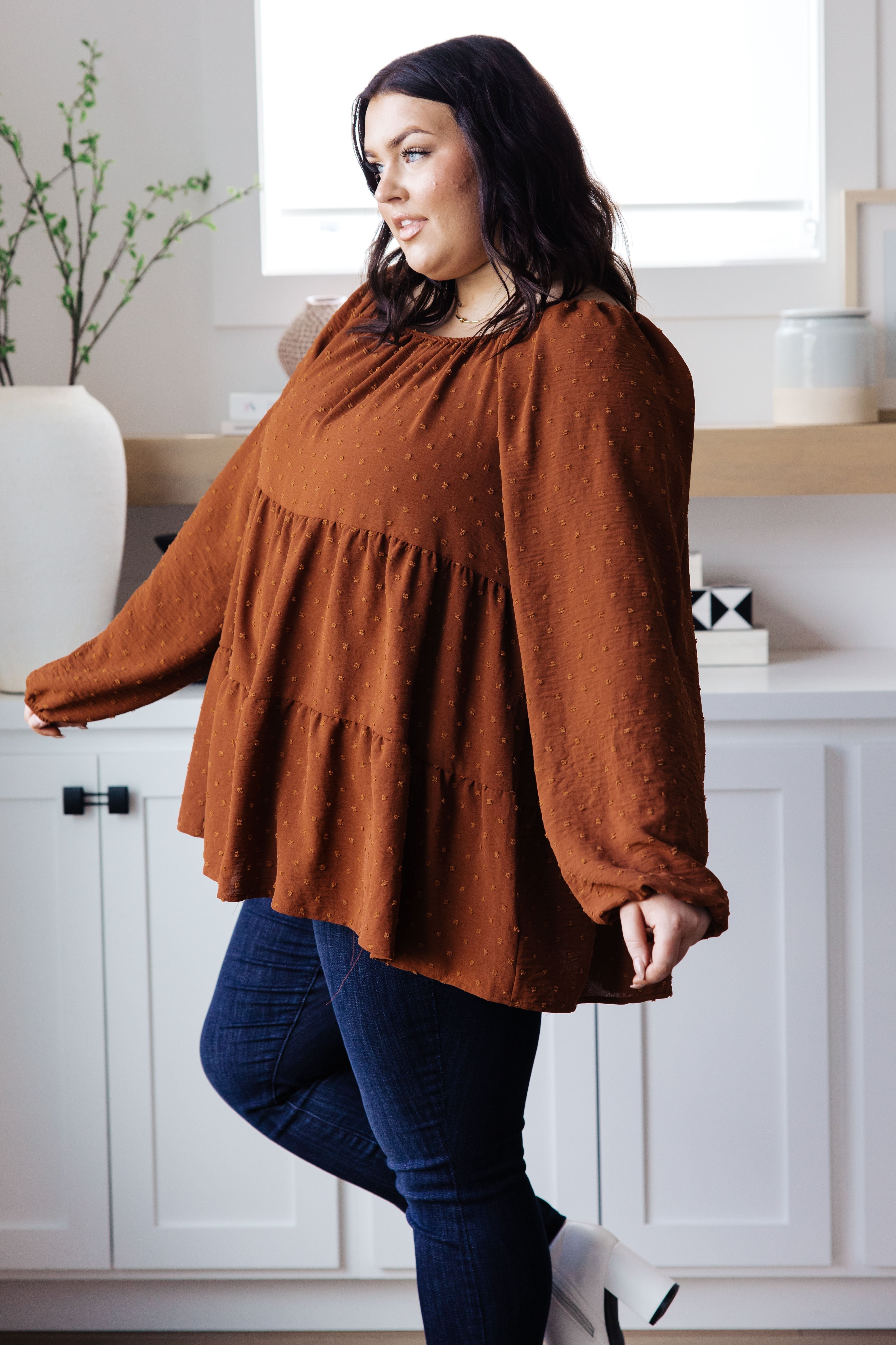 Plus-size woman wearing rust tiered babydoll top with long puffed sleeves and dark blue jeans indoors