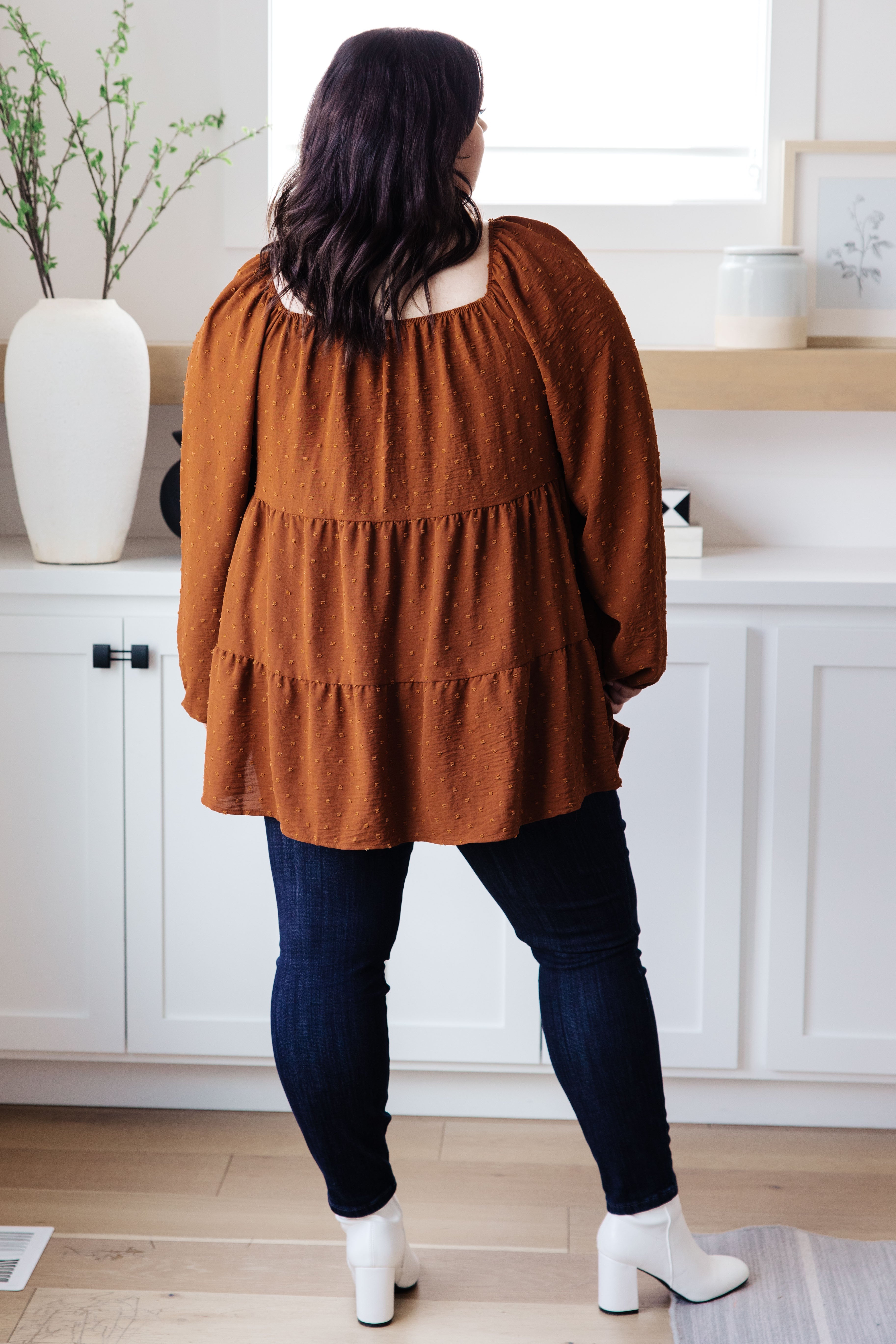 Woman wearing rust tiered babydoll top with long sleeves, paired with dark skinny jeans and white ankle boots indoors
