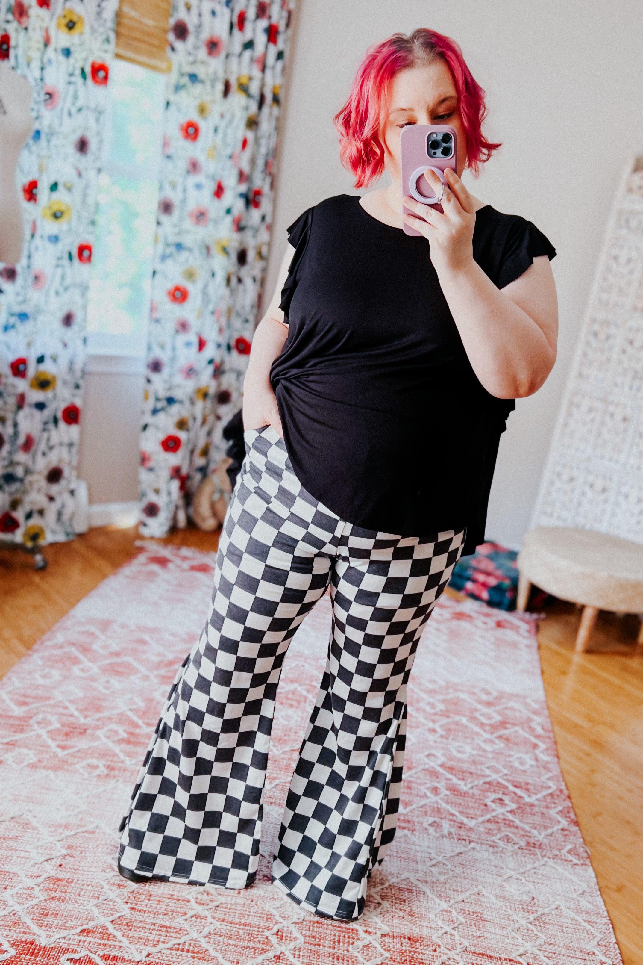 Plus-size woman wearing black and white checkered flared pants with a black top taking mirror selfie indoors