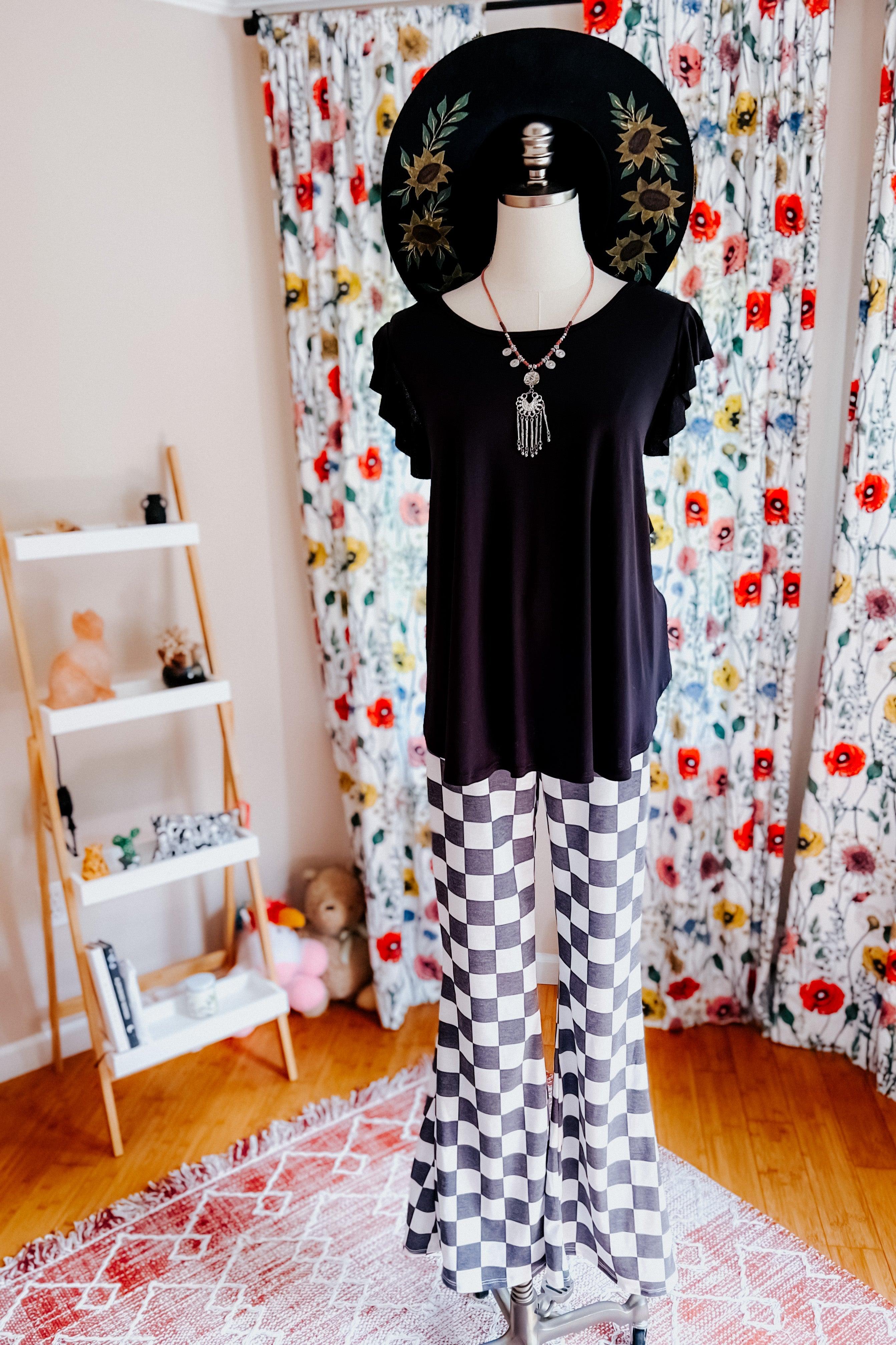 Mannequin dressed in black top, checkered flared pants, and black wide-brim hat with floral design in cozy room setting