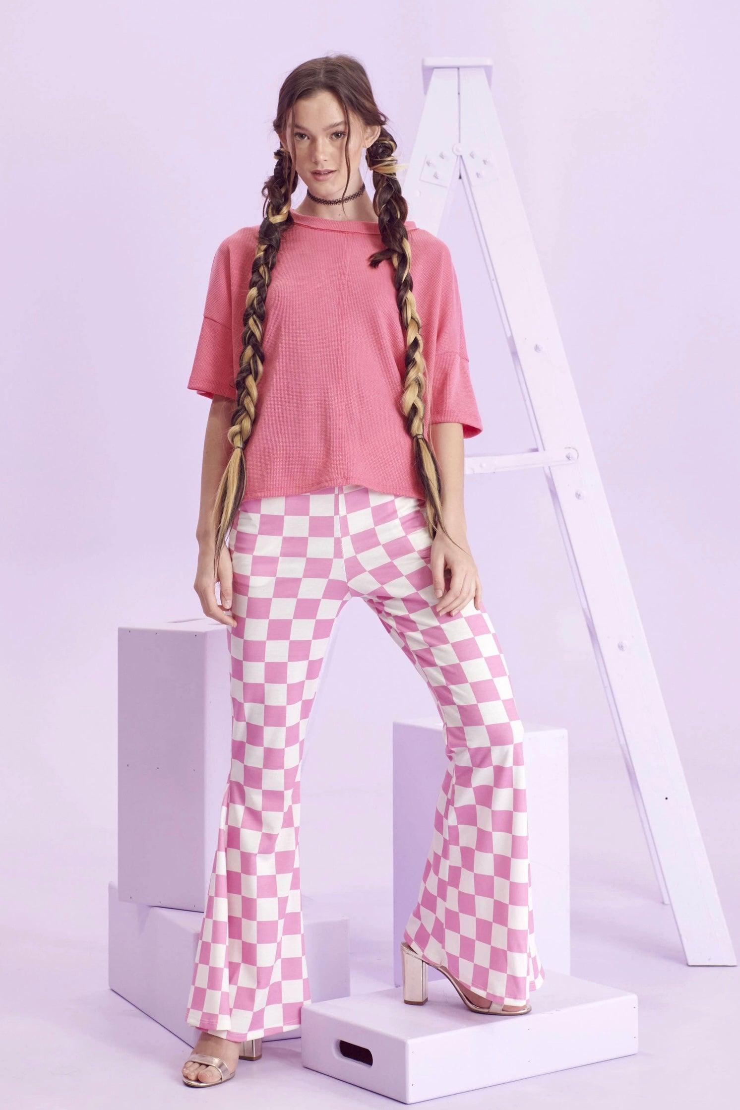 Model wearing pink and white checkerboard flared pants paired with a loose pink short-sleeve top standing on platform blocks