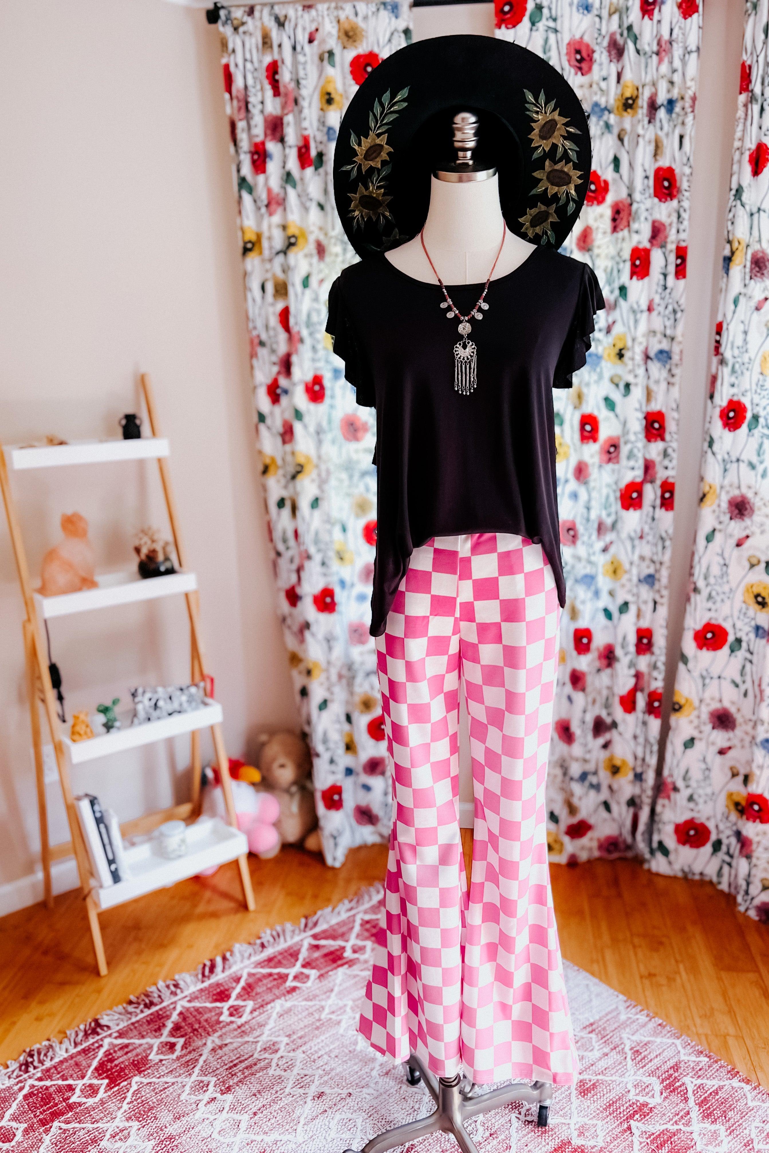 Mannequin dressed in pink and white checkered flared pants with a black top and wide-brimmed hat in floral room setting