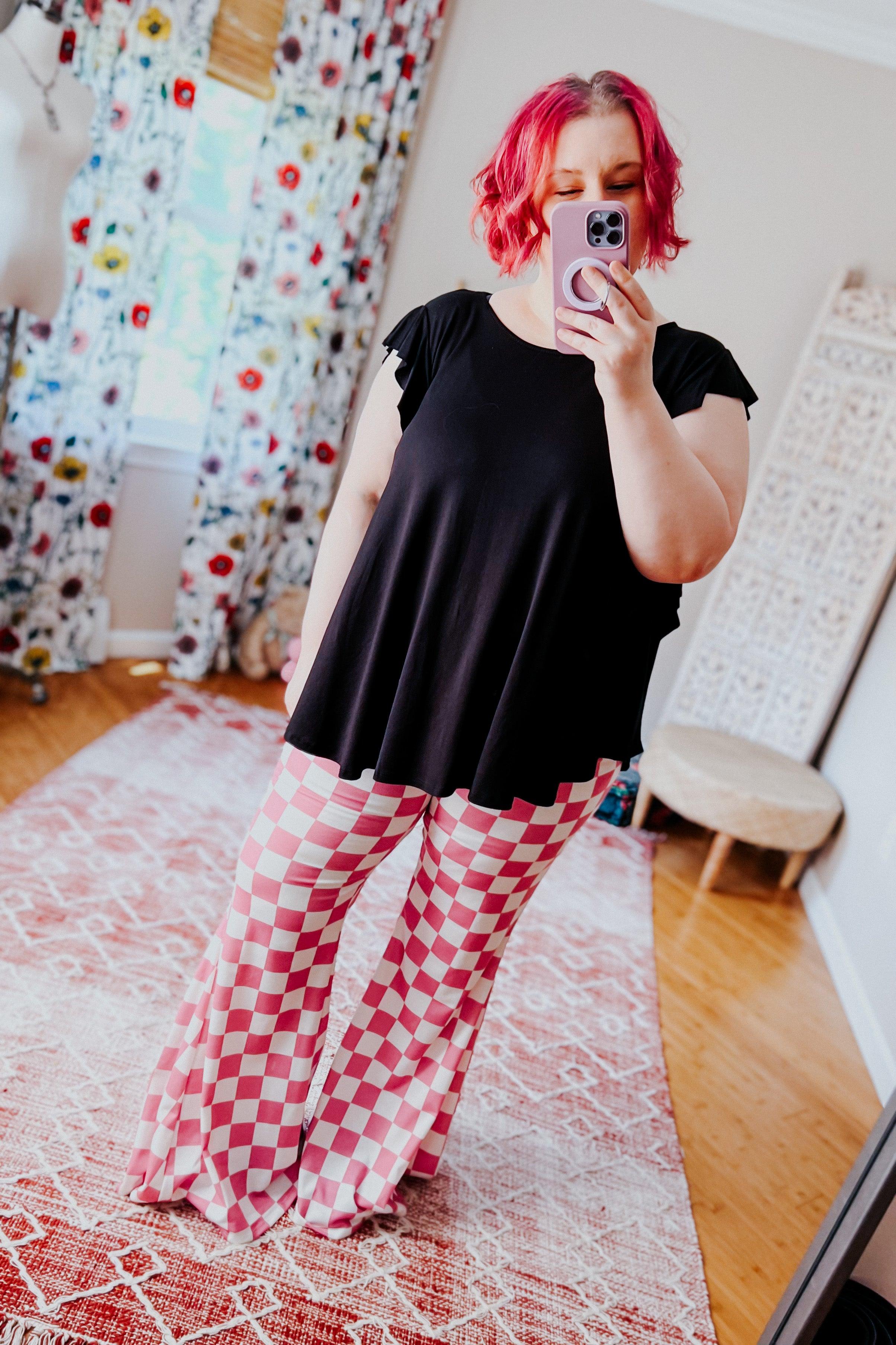 Woman wearing pink and white checkered flared pants and black top taking a selfie in a brightly lit room with floral curtains