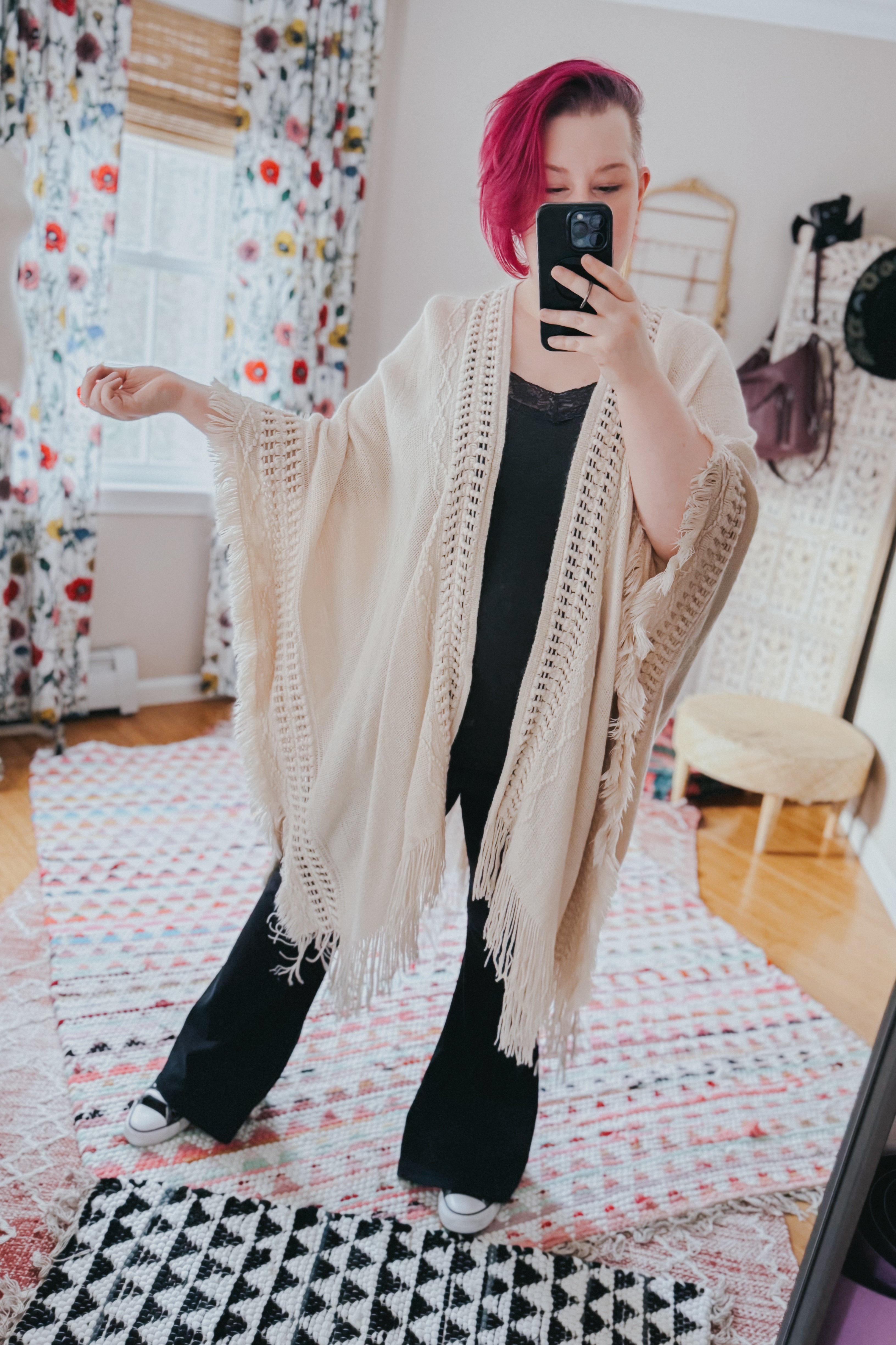 Woman with magenta hair wearing a cream boho ruana with fringed edges over black outfit taking selfie indoors