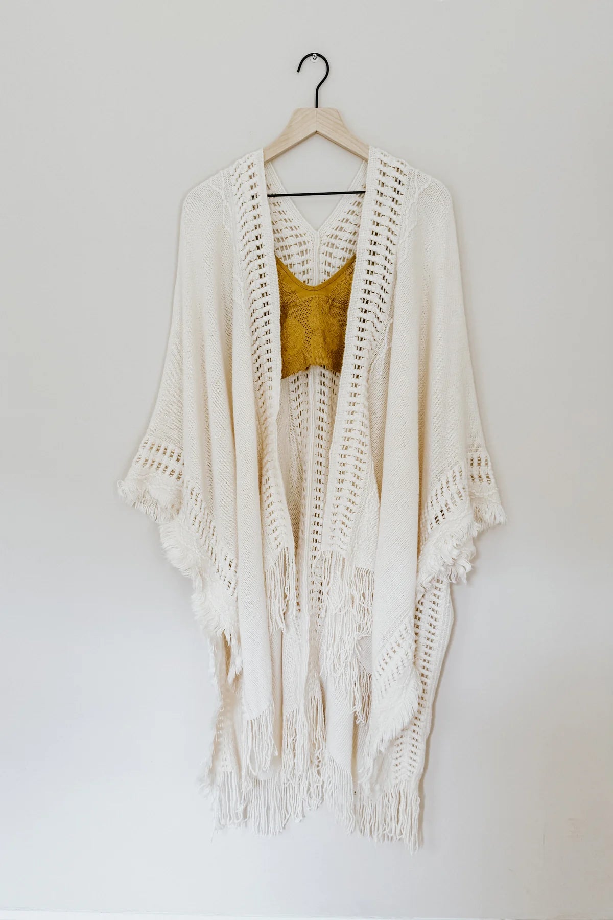 Cream bohemian ruana with intricate open knit panels and fringed hem displayed on wooden hanger with mustard knit top inside