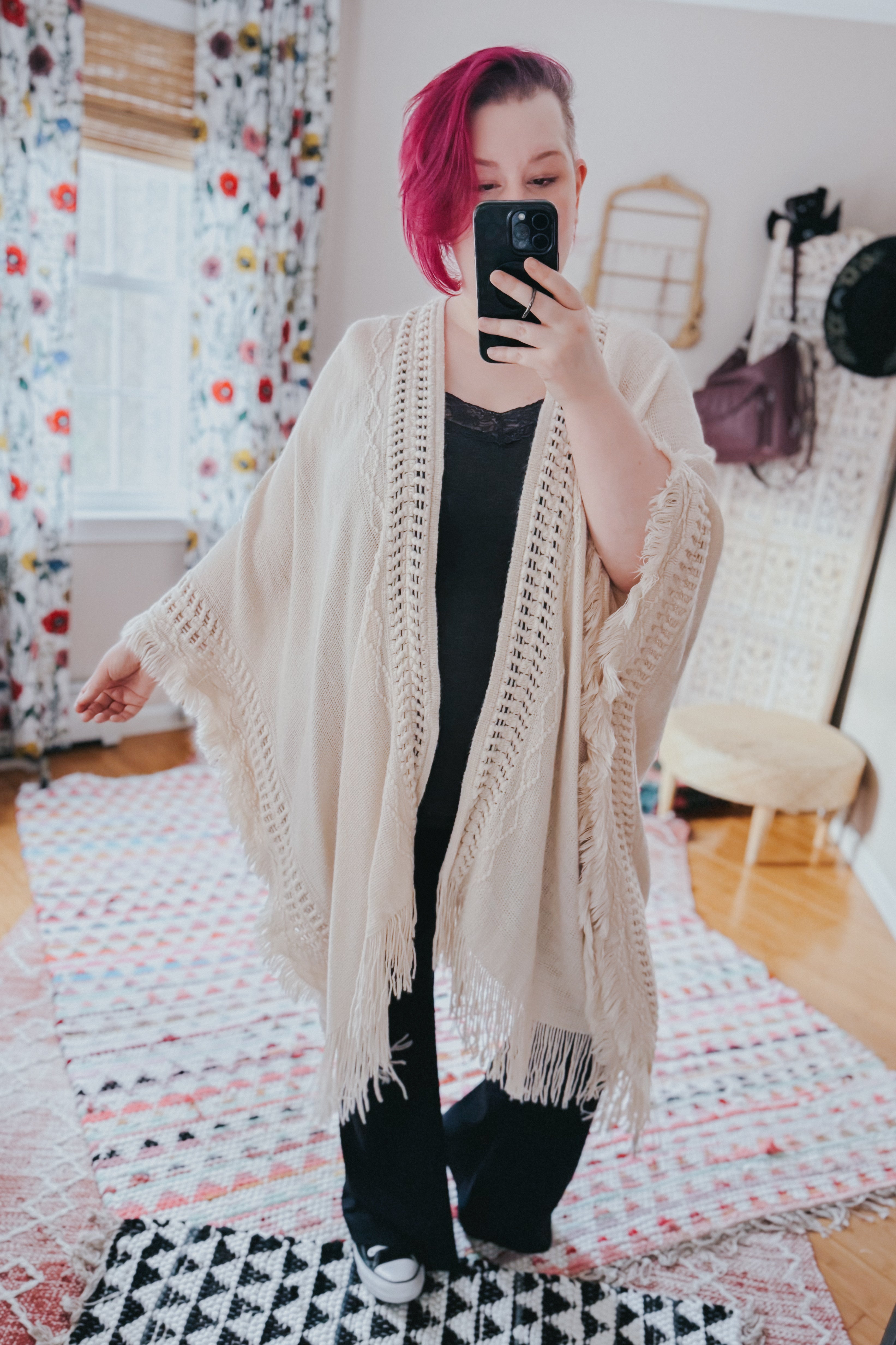 Woman with vibrant magenta hair wearing a cream bohemian ruana with frayed edges taking a mirror selfie indoors