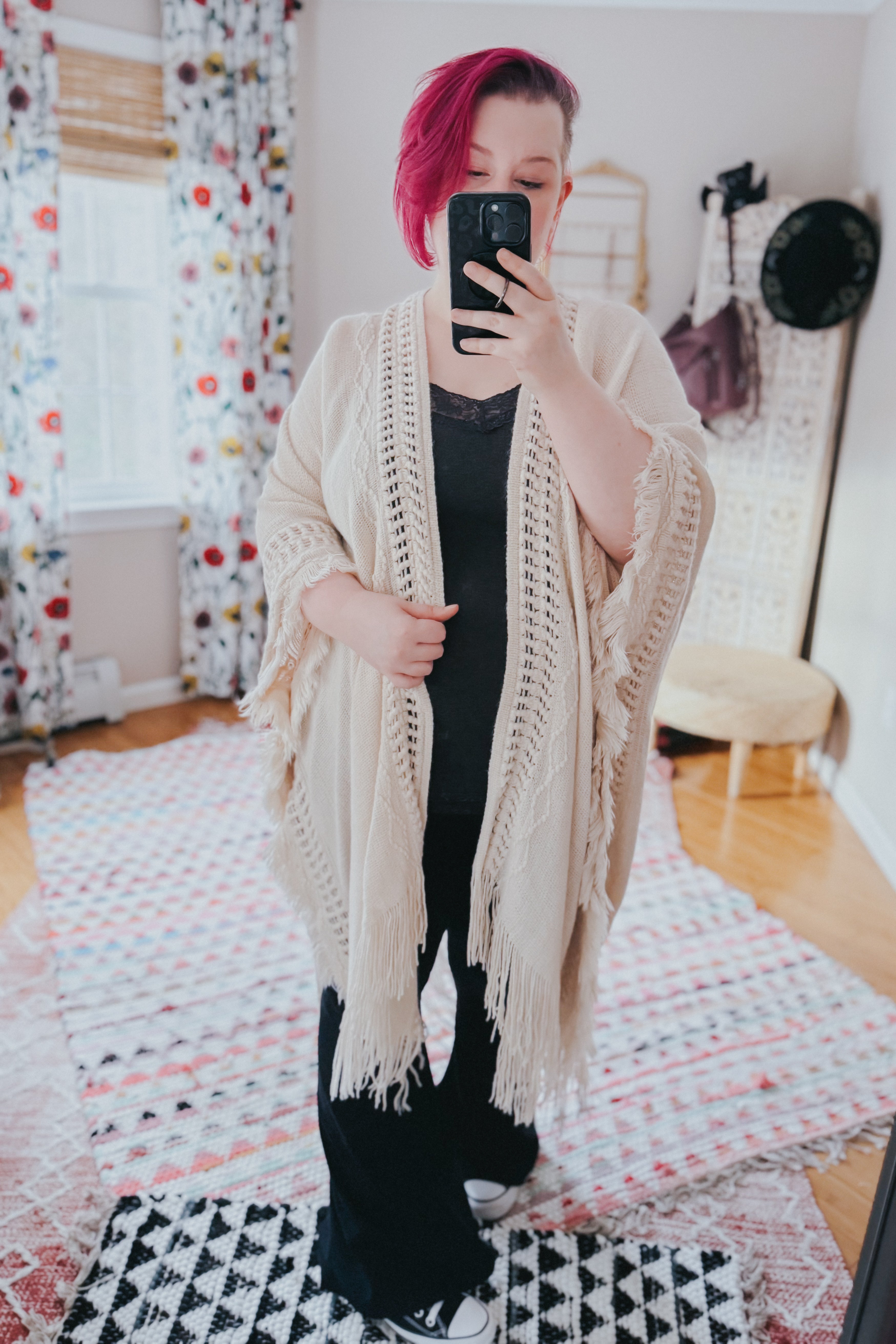 Woman with short magenta hair wearing a cream bohemian ruana with fringed and textured borders taking a mirror selfie indoors
