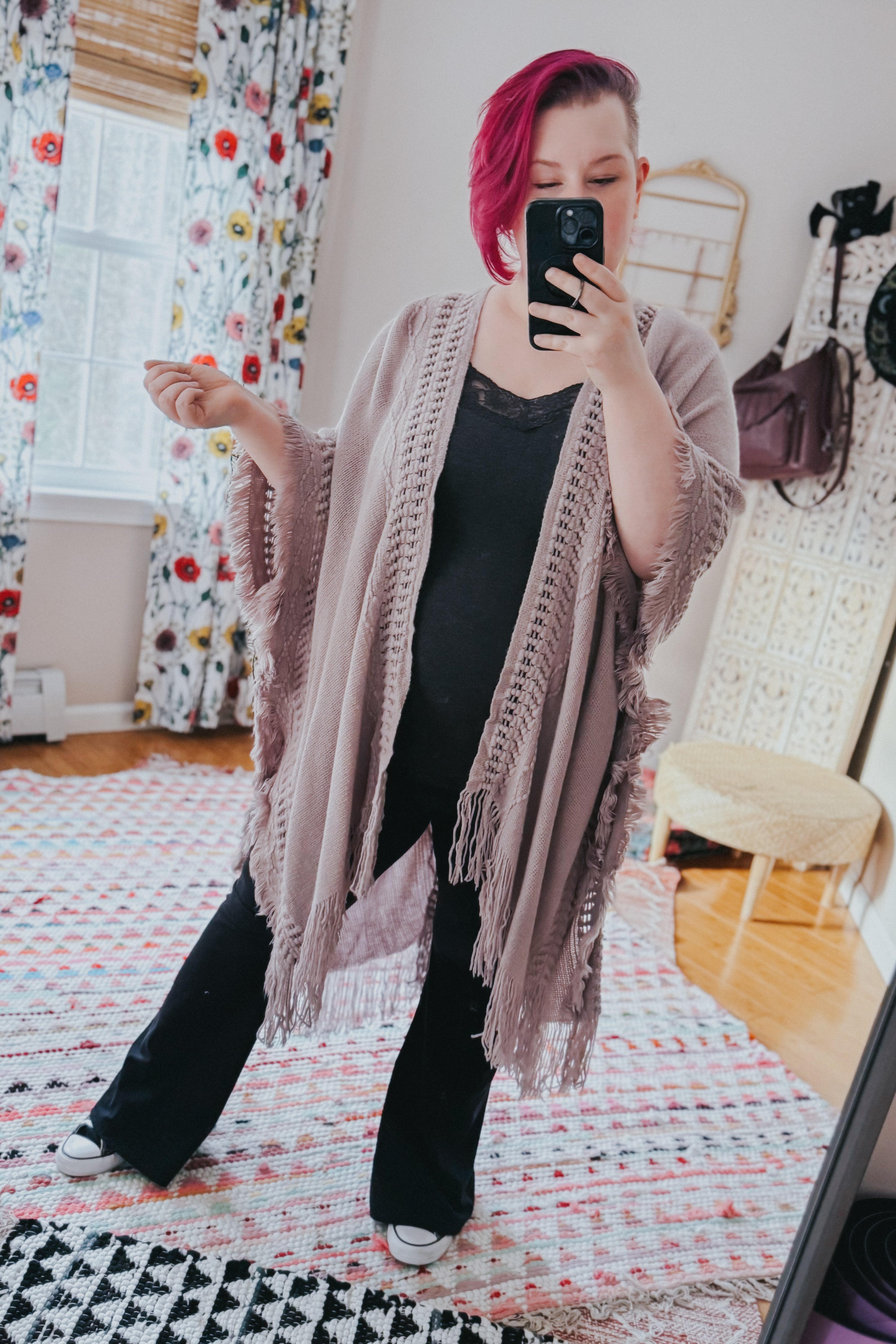 Woman with magenta hair wearing a grey bohemian ruana with open knit panels and fringed edges taking indoor mirror selfie