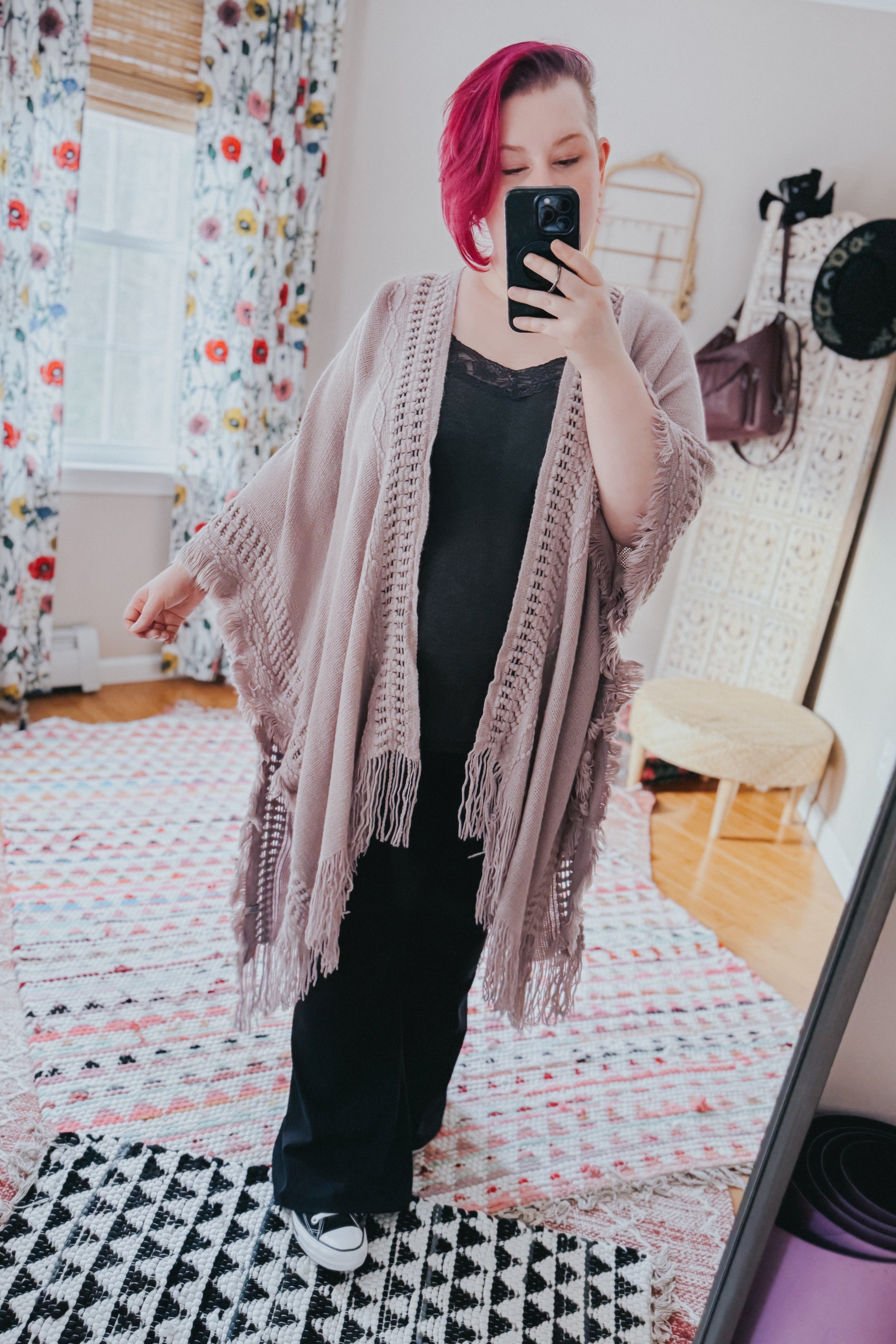 Woman with short magenta hair wearing a grey bohemian ruana with frayed edges and fringed details taking a mirror selfie indoors