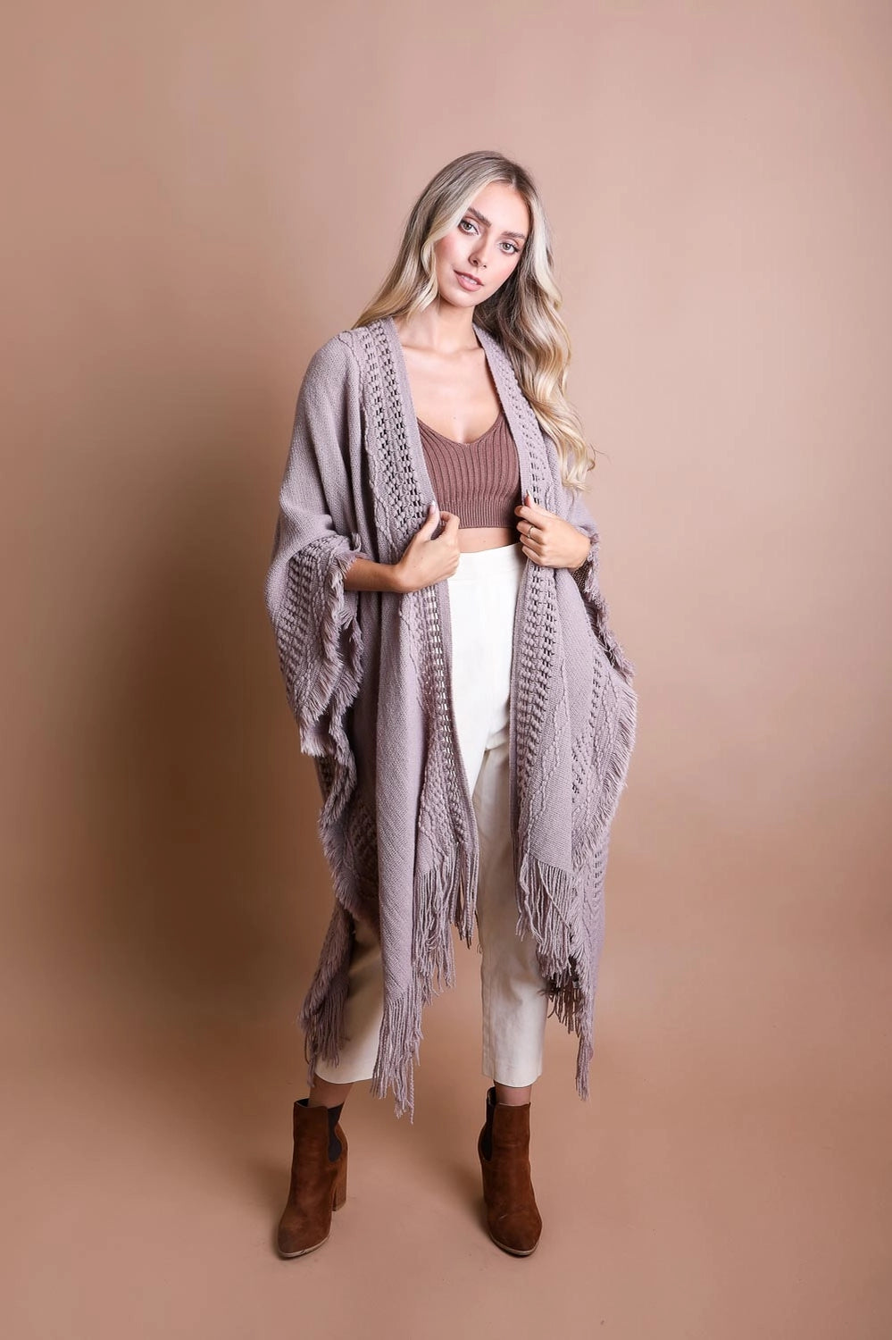 Blonde woman wearing a taupe knitted ruana with frayed edges and fringe, styled with brown top and white pants standing against beige backdrop