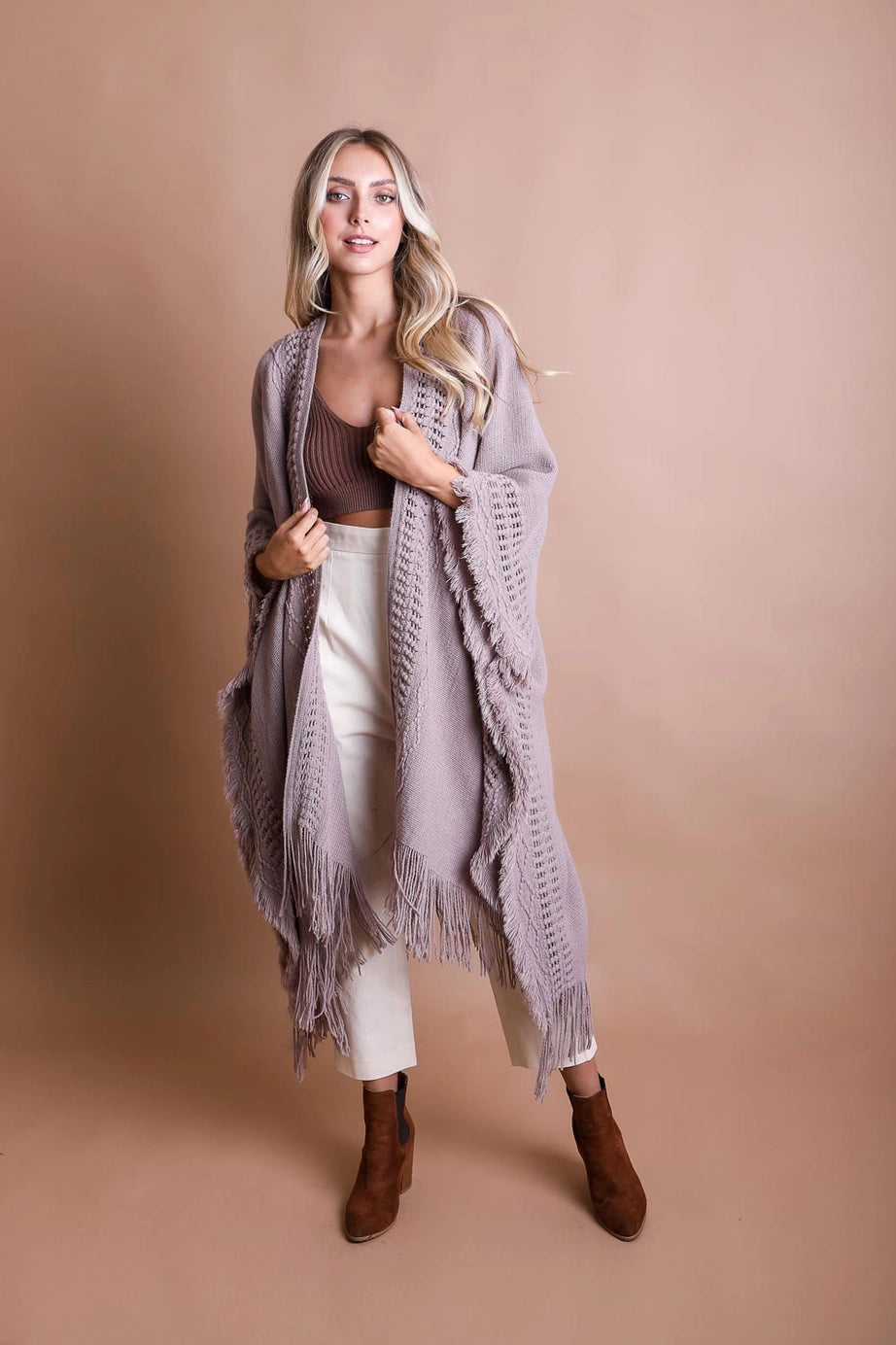Blonde woman wearing a grey boho ruana with frayed edges and long fringe, styled with brown top and cream pants against brown backdrop
