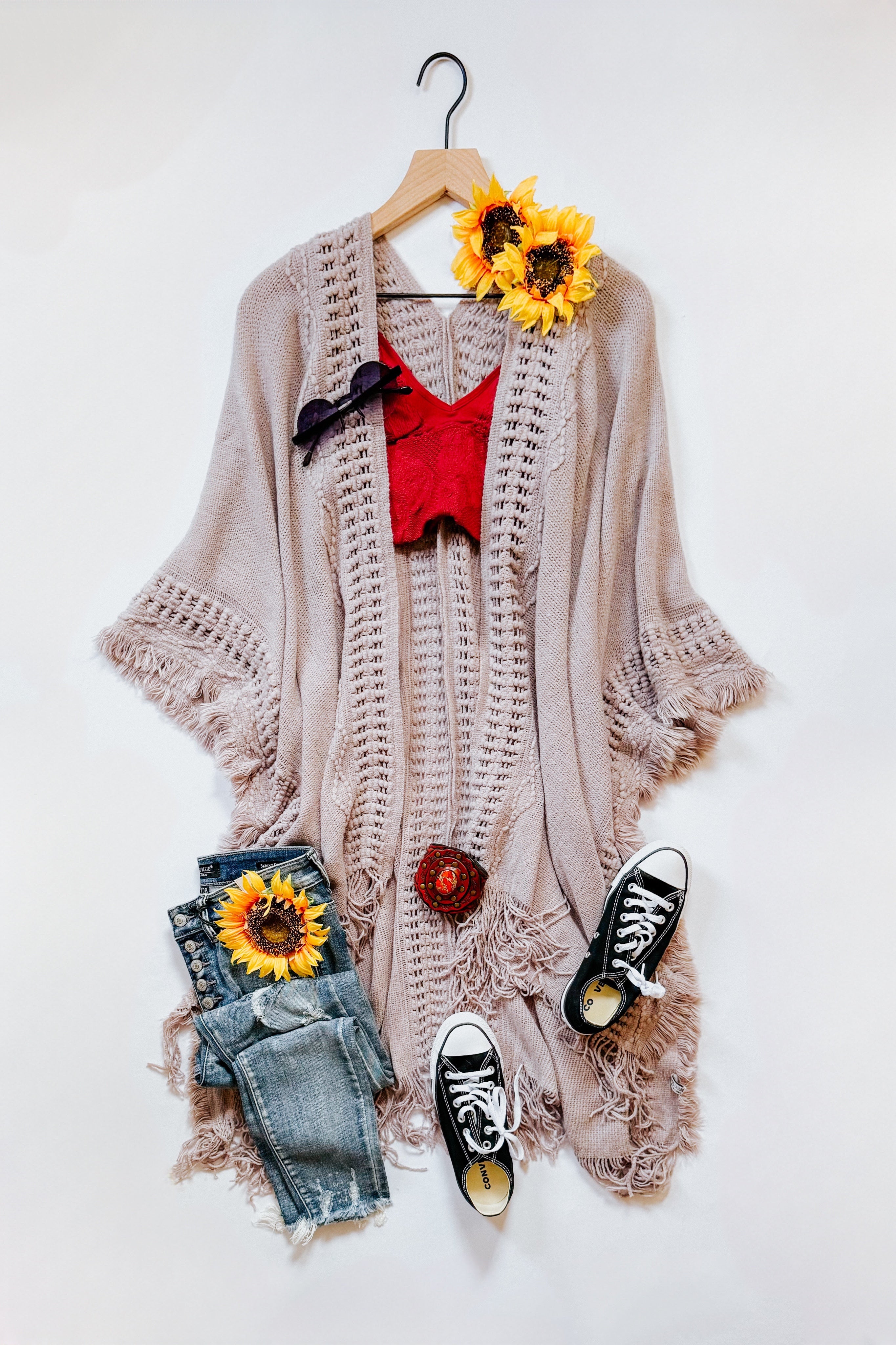 Grey knitted bohemian ruana with fringed edges styled with red crop top, distressed jeans, and black sneakers flat lay with sunflowers
