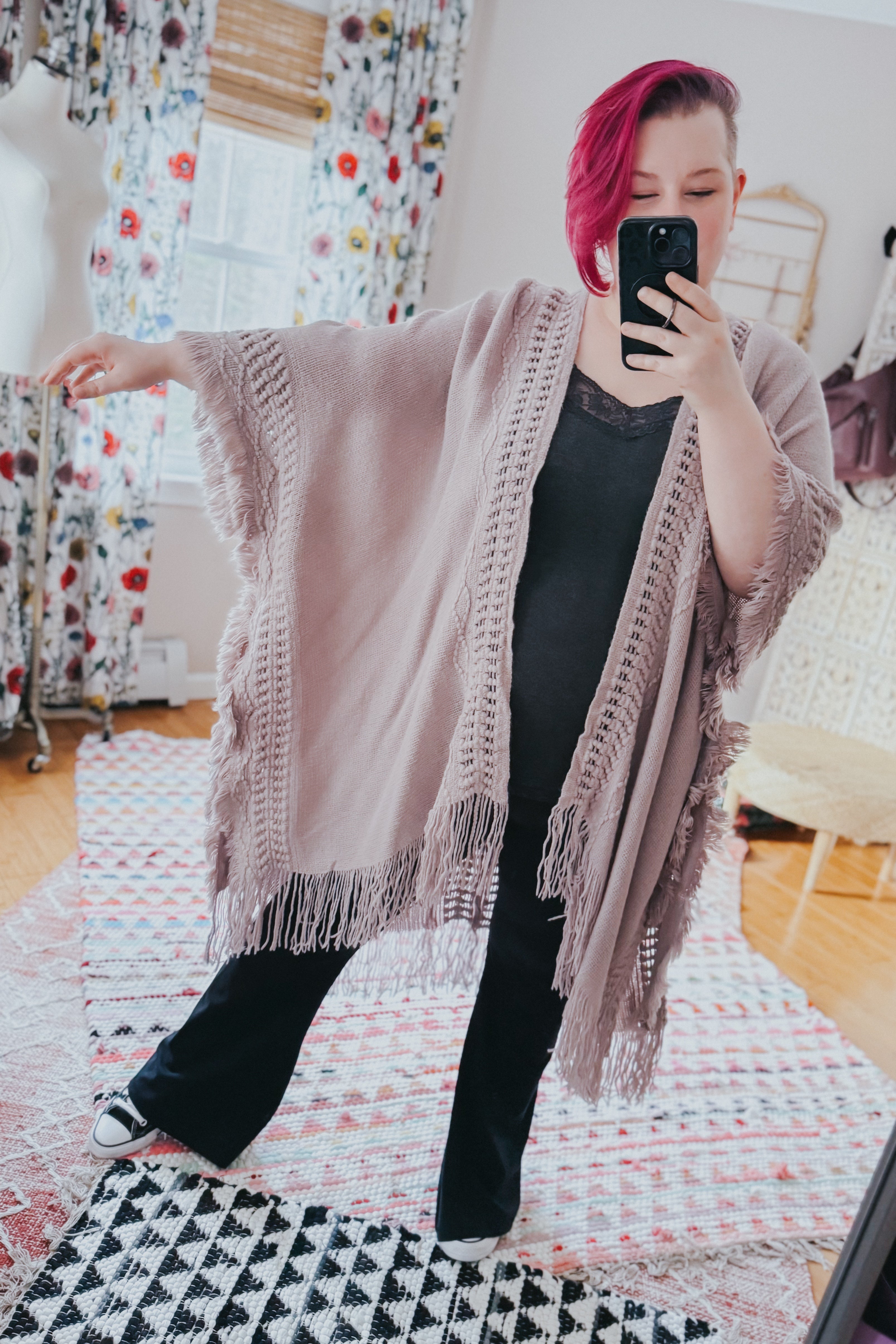 Woman with bright magenta hair wearing a soft grey ruana with open knit trim and long fringed edges taking a mirror selfie indoors