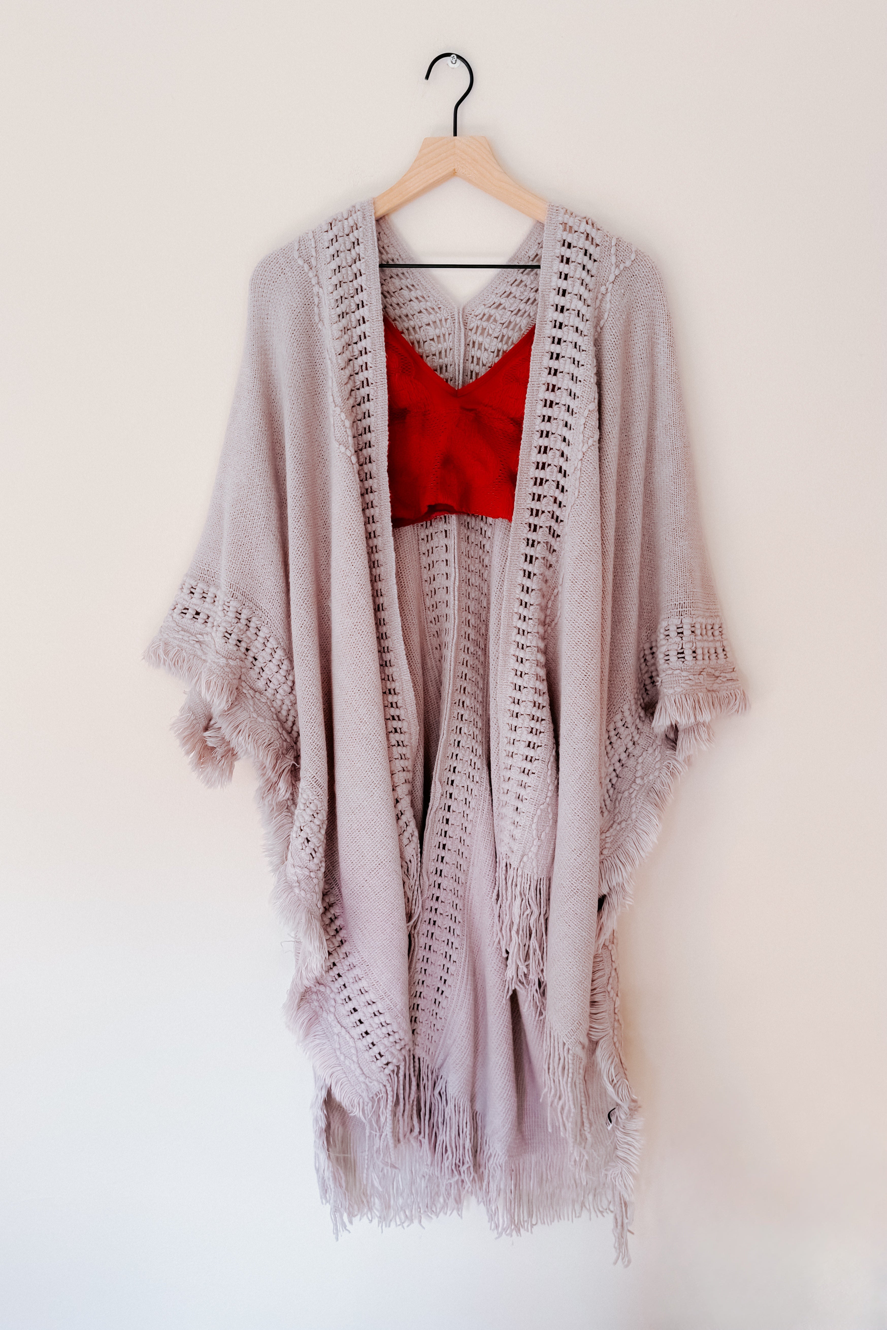 Grey knitted bohemian ruana with open stitch details and long fringed hem hanging on wooden hanger against white wall