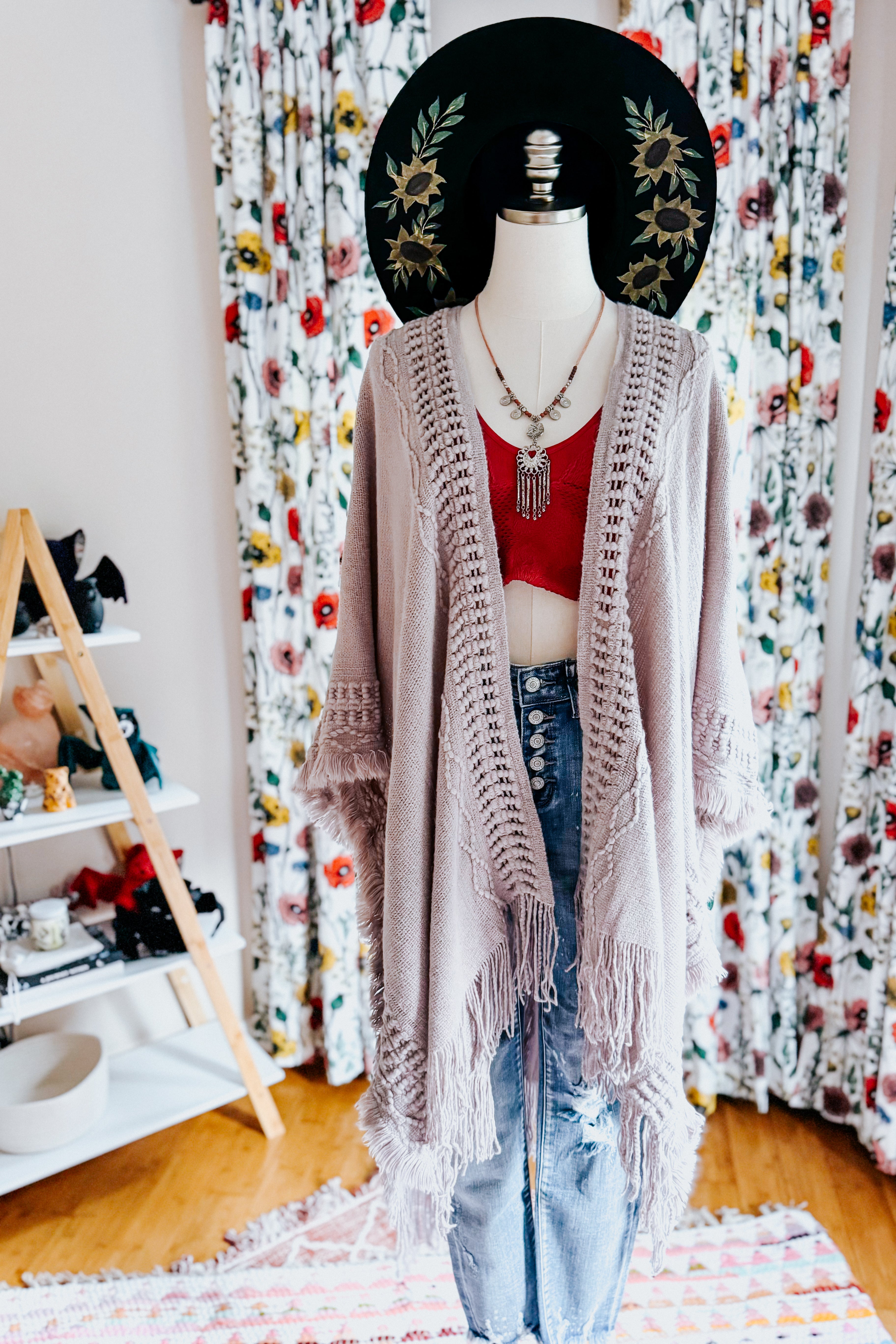 Mannequin dressed in a soft grey fringed ruana layered over red crop top and high-waisted button fly jeans with floral backdrop