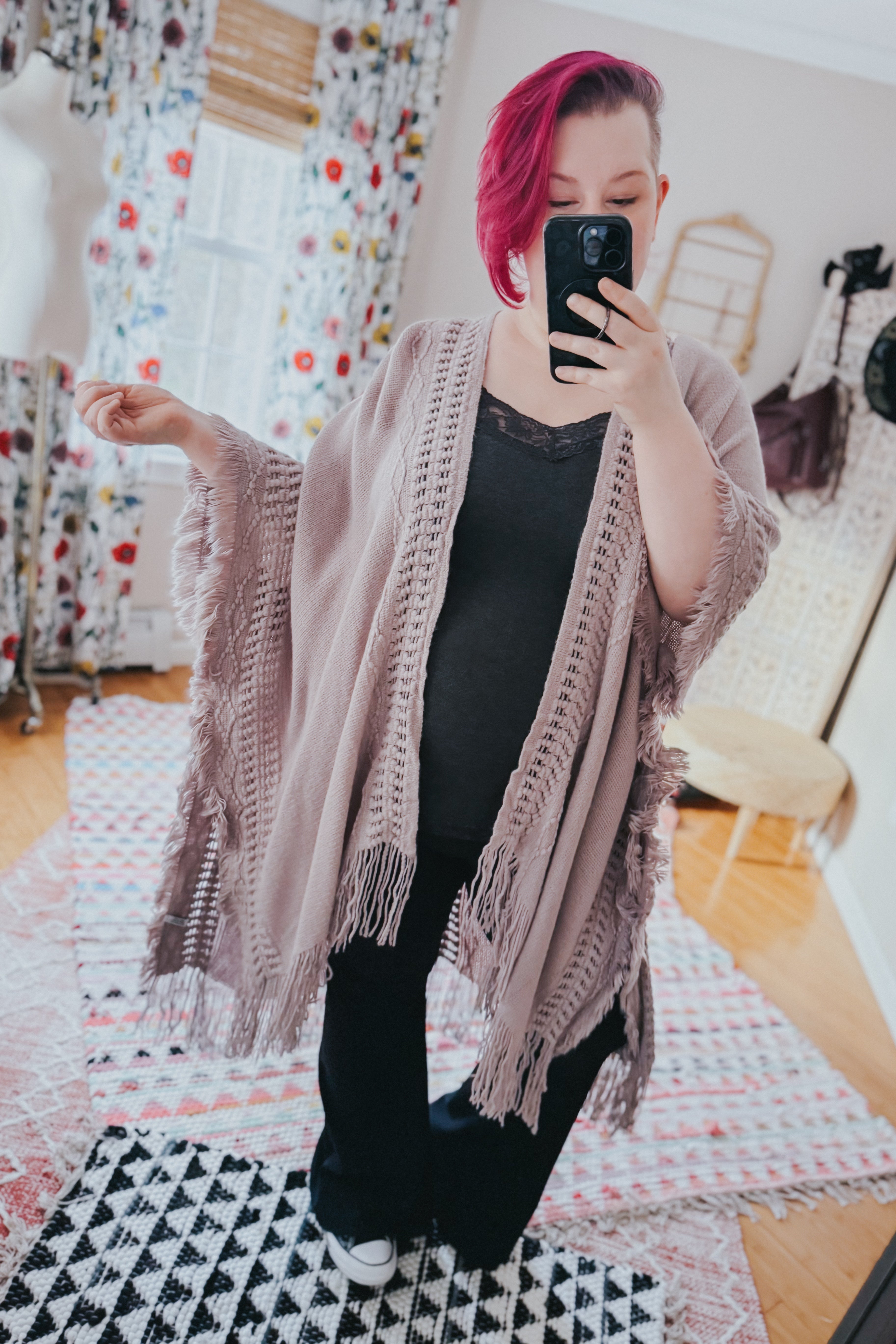 Woman with vibrant magenta hair wearing a grey ruana with textured borders and fringe taking a mirror selfie indoors