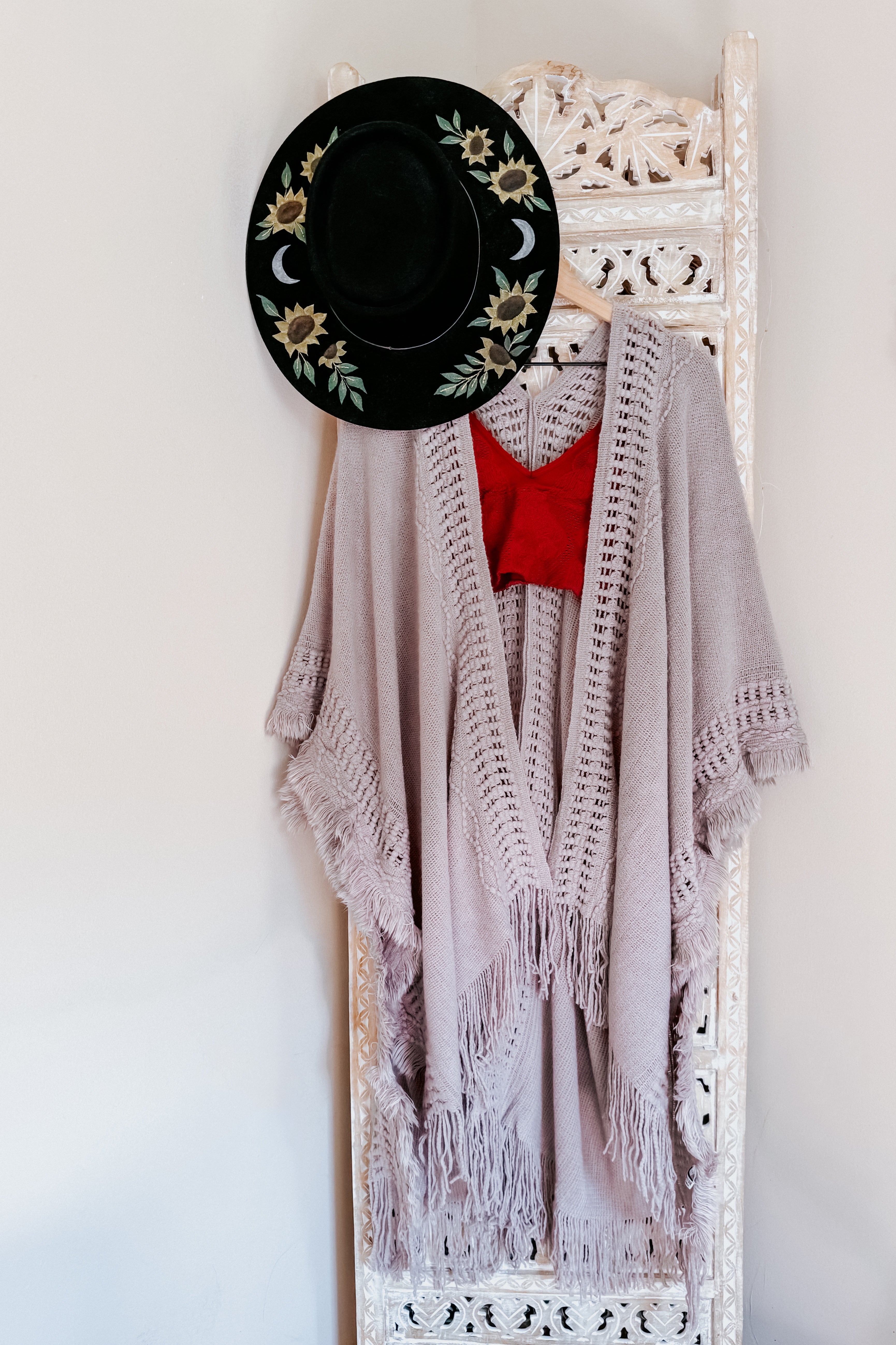 Grey bohemian ruana with frayed edges and open knit details draped on carved wooden screen with black floral hat