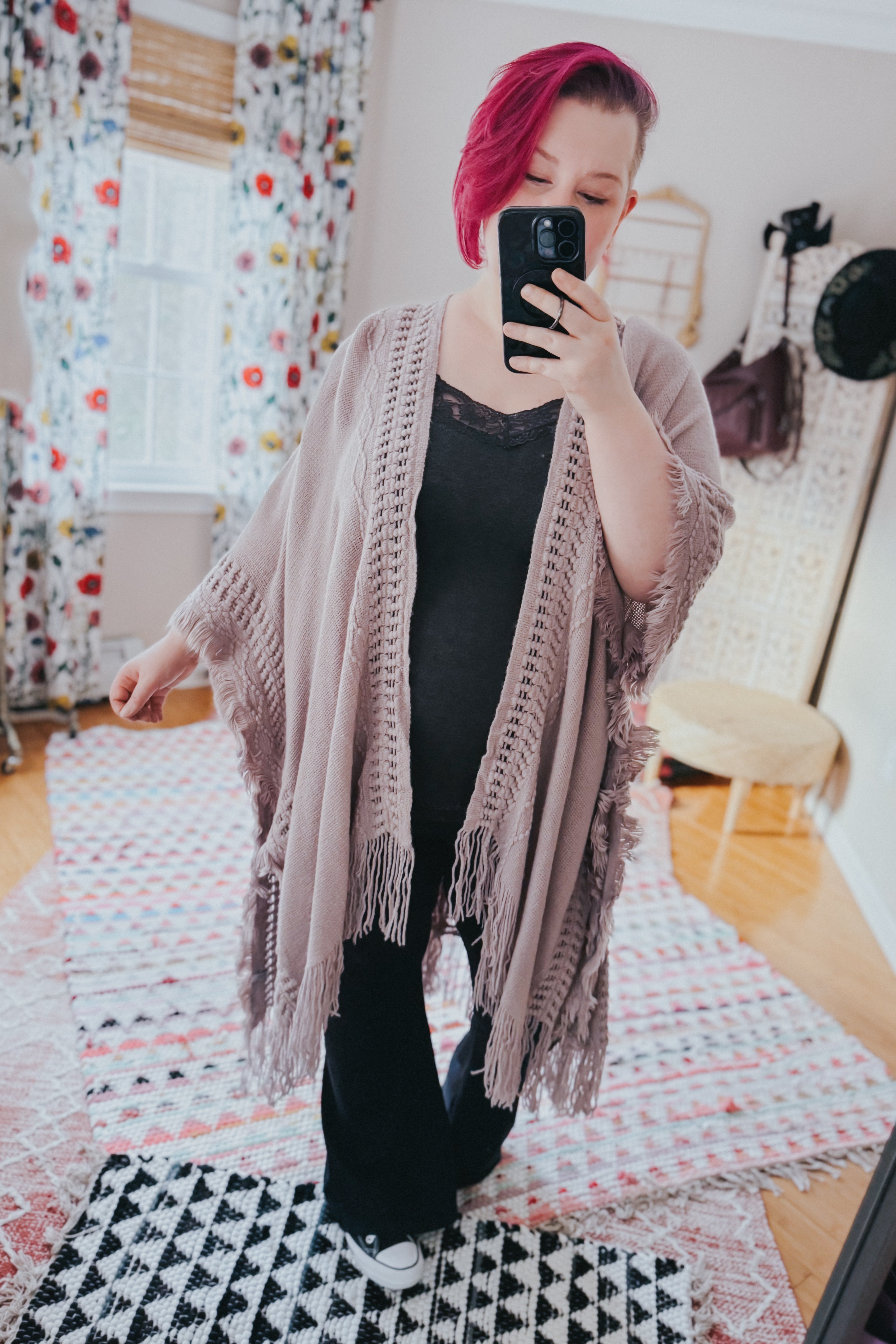 Woman with magenta hair wearing a soft grey knitted ruana with fringed edges taking a mirror selfie indoors