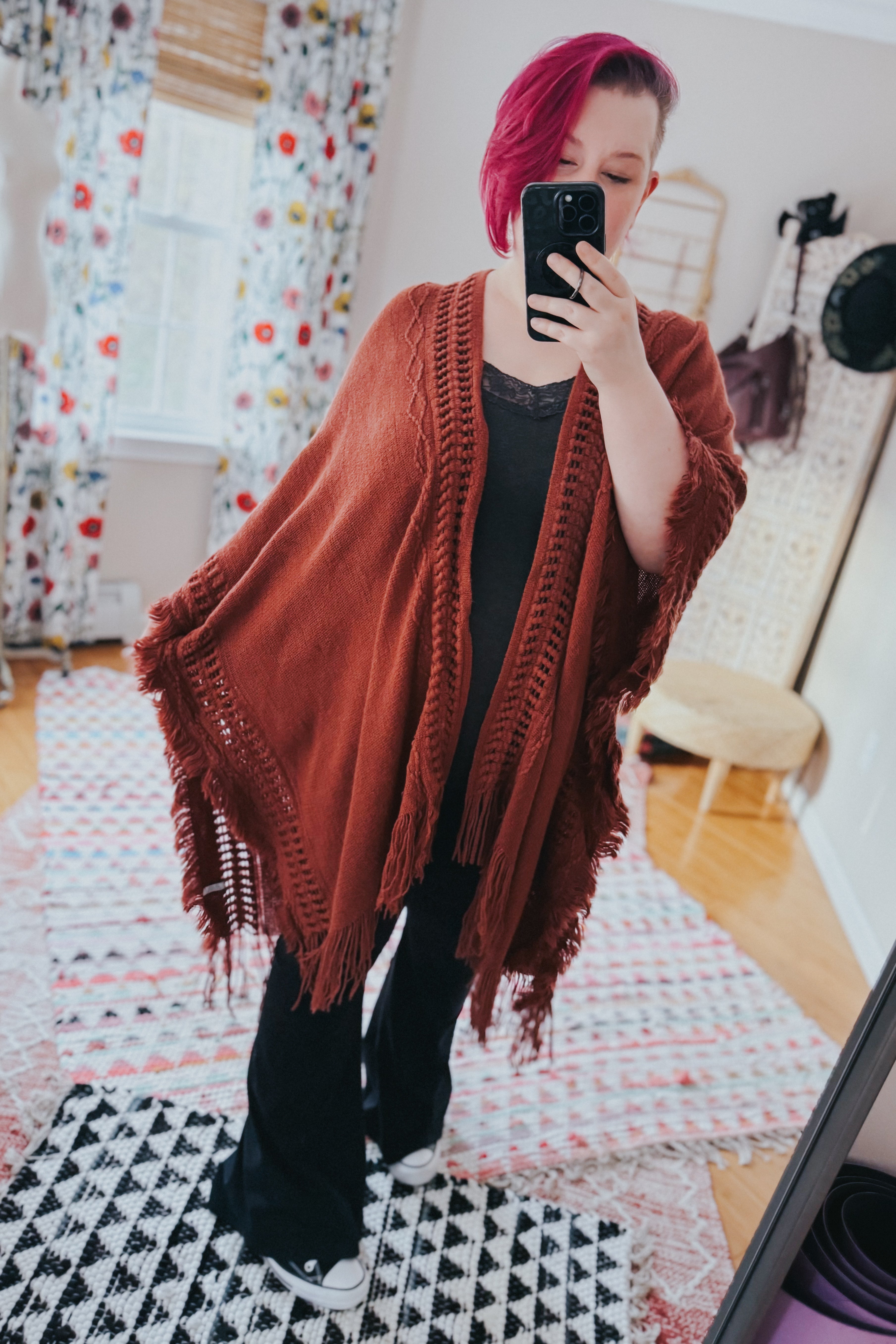 Woman wearing a rust-colored bohemian fringed ruana over black clothing, taking a mirror selfie in a cozy room