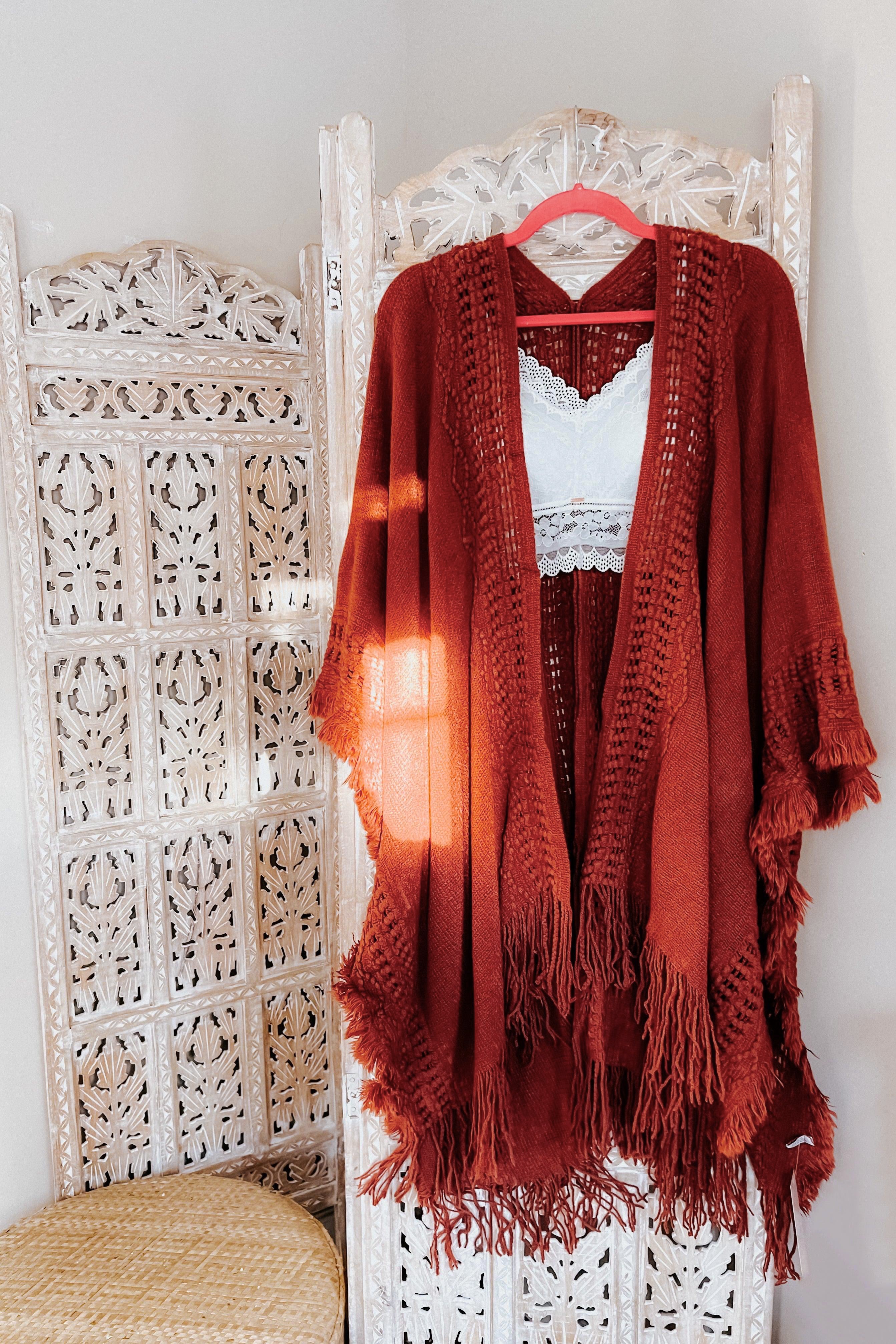 Rust-colored bohemian ruana with frayed edges layered over white lace bralette on carved wooden screen.