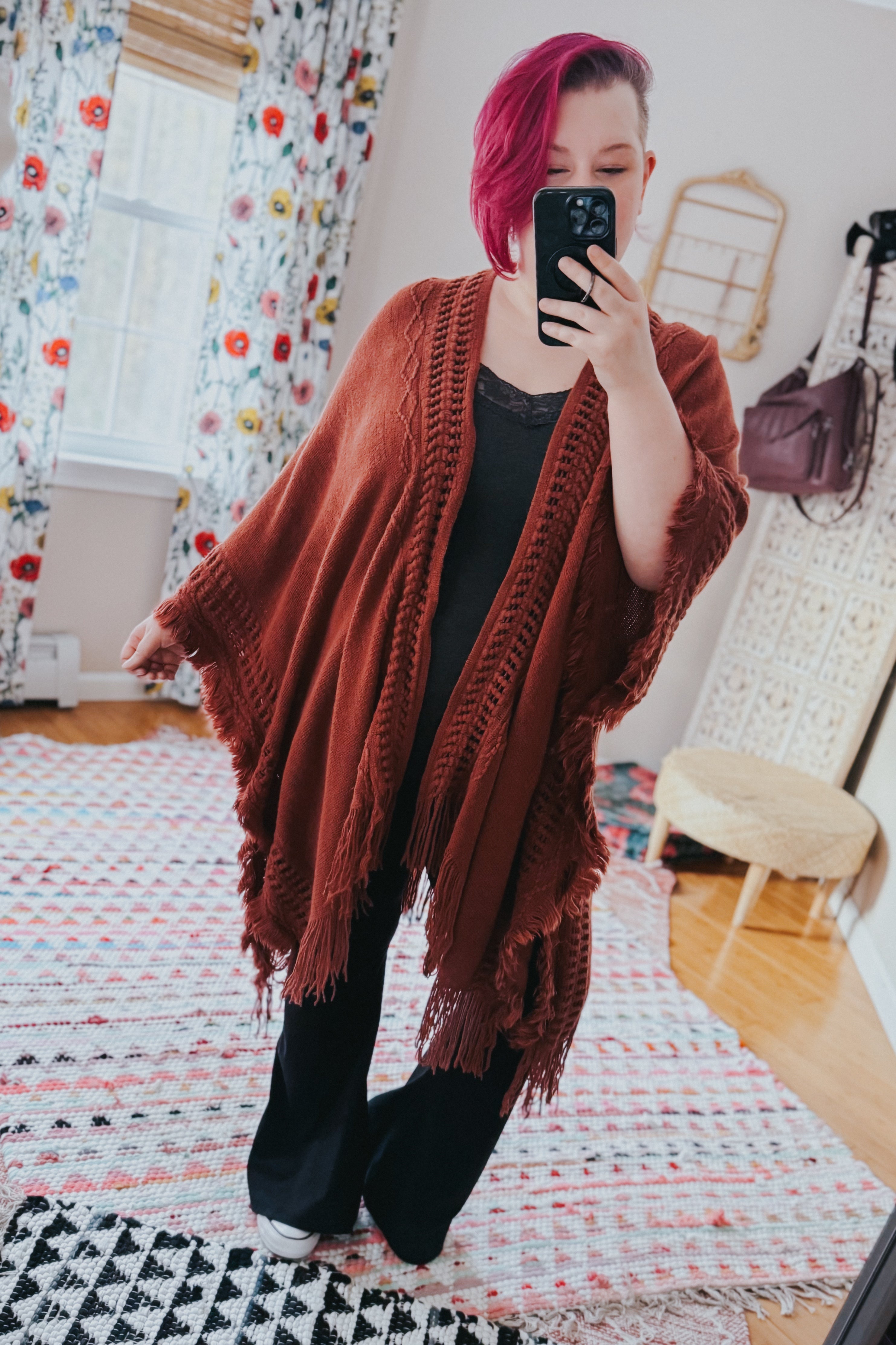 Woman wearing a rust-colored bohemian ruana with fringe and textured knit standing in a bright, eclectic room