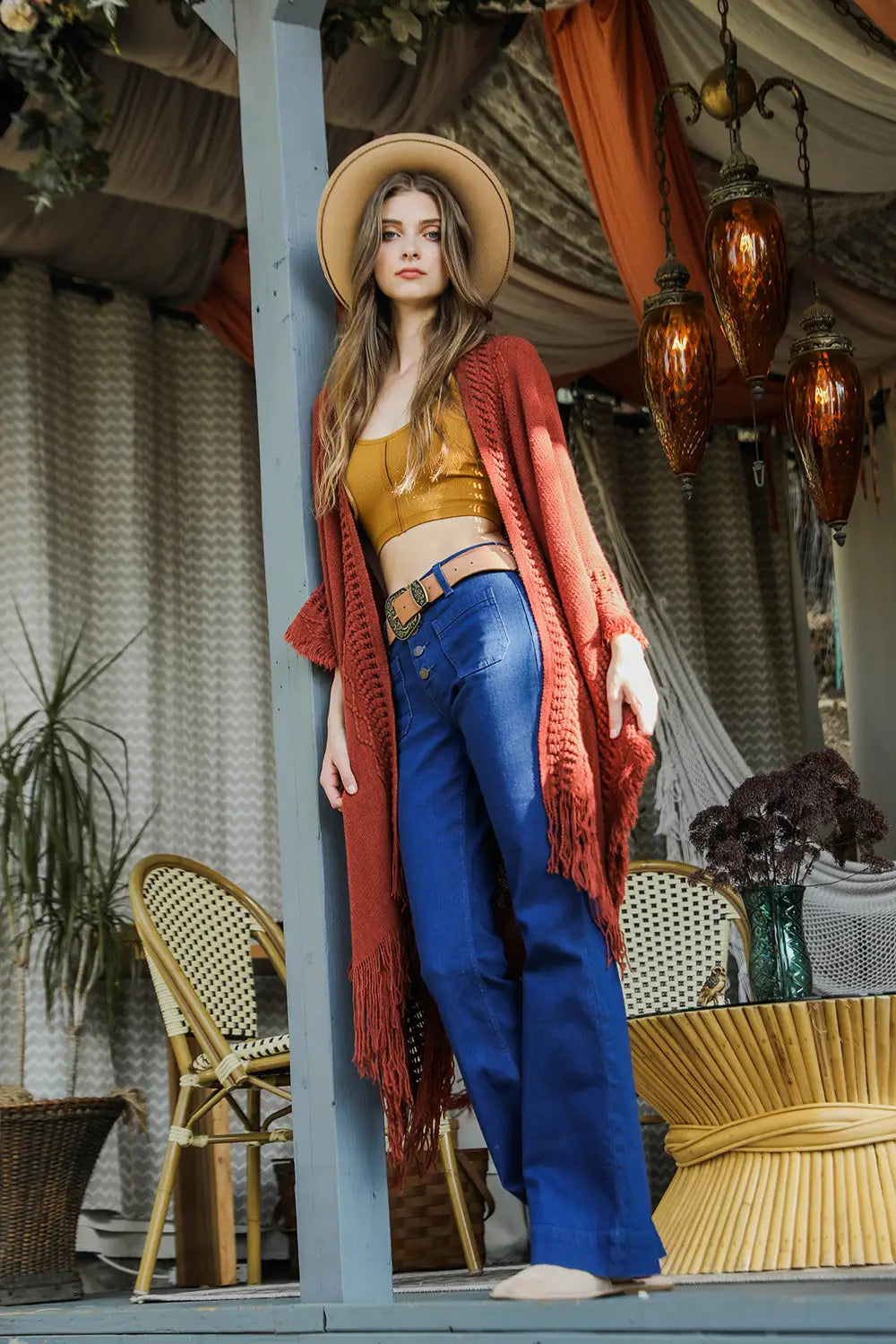 Woman wearing a rust-colored fringed bohemian ruana over a mustard crop top with high-waisted blue pants outdoors