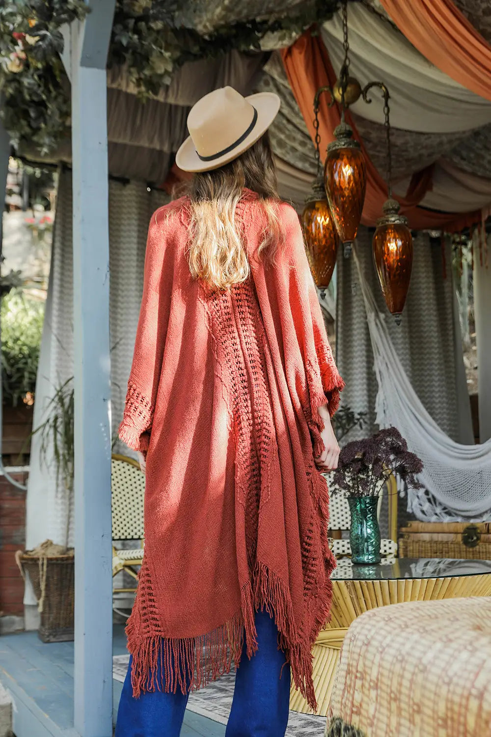 Woman wearing a rust-colored bohemian ruana with fringe and a wide-brimmed hat in a cozy indoor setting