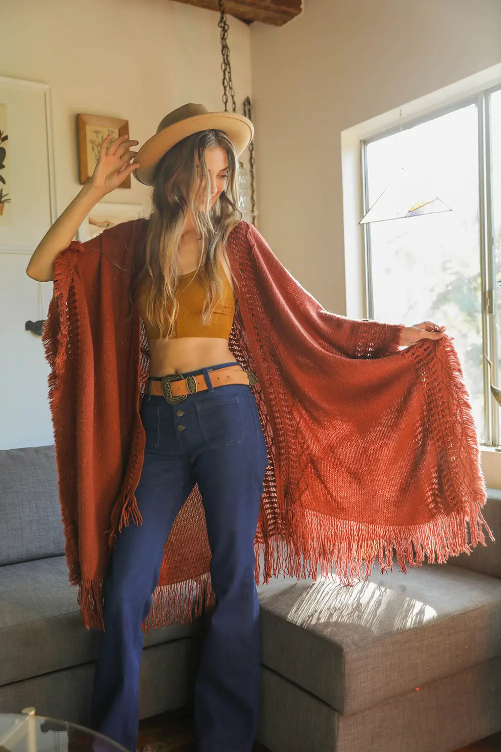 Woman wearing a rust-colored fringed bohemian ruana with a tank top, high-waisted jeans, and a wide-brim hat indoors