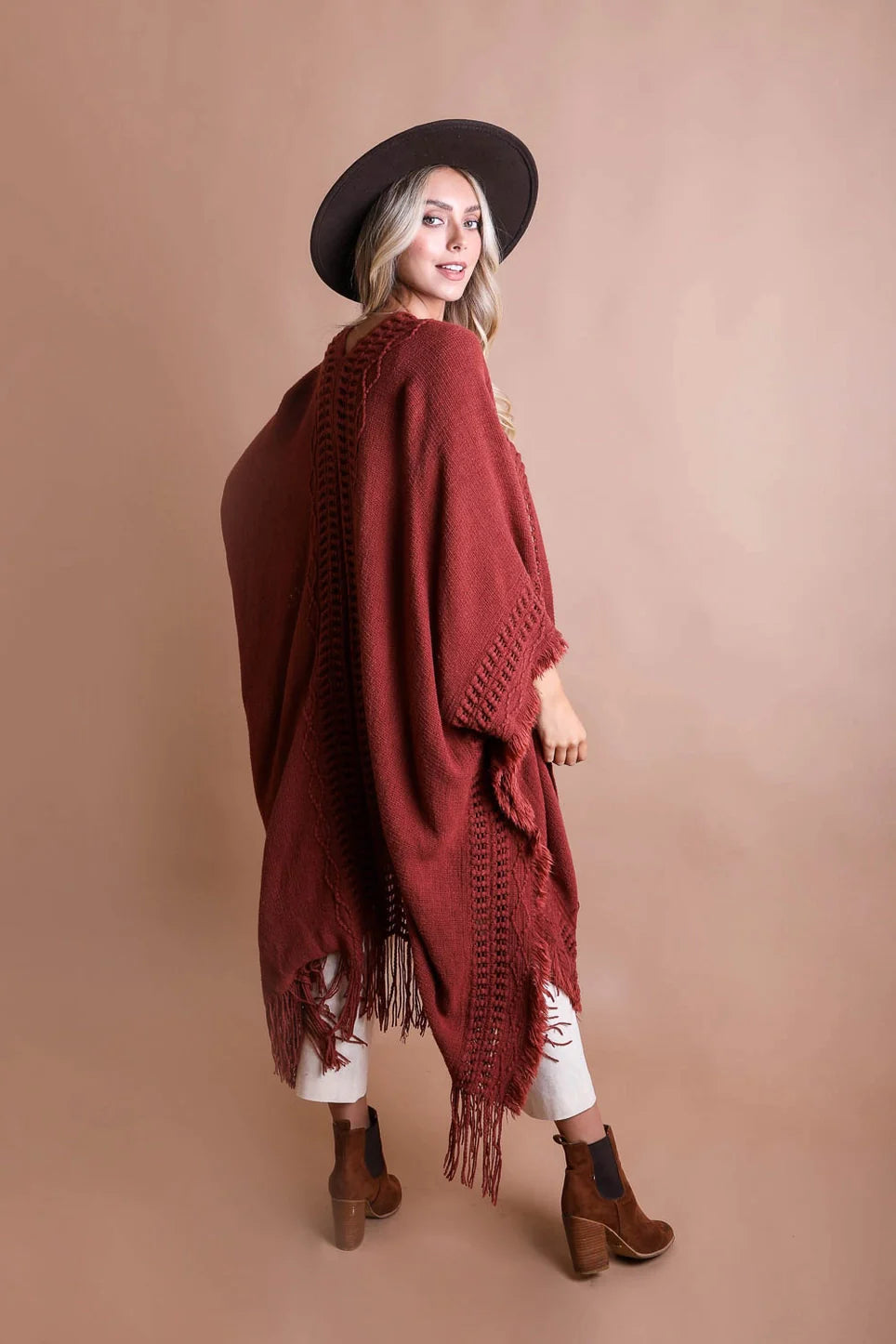 Woman wearing a rust-colored bohemian ruana with frayed edges paired with white pants and brown ankle boots