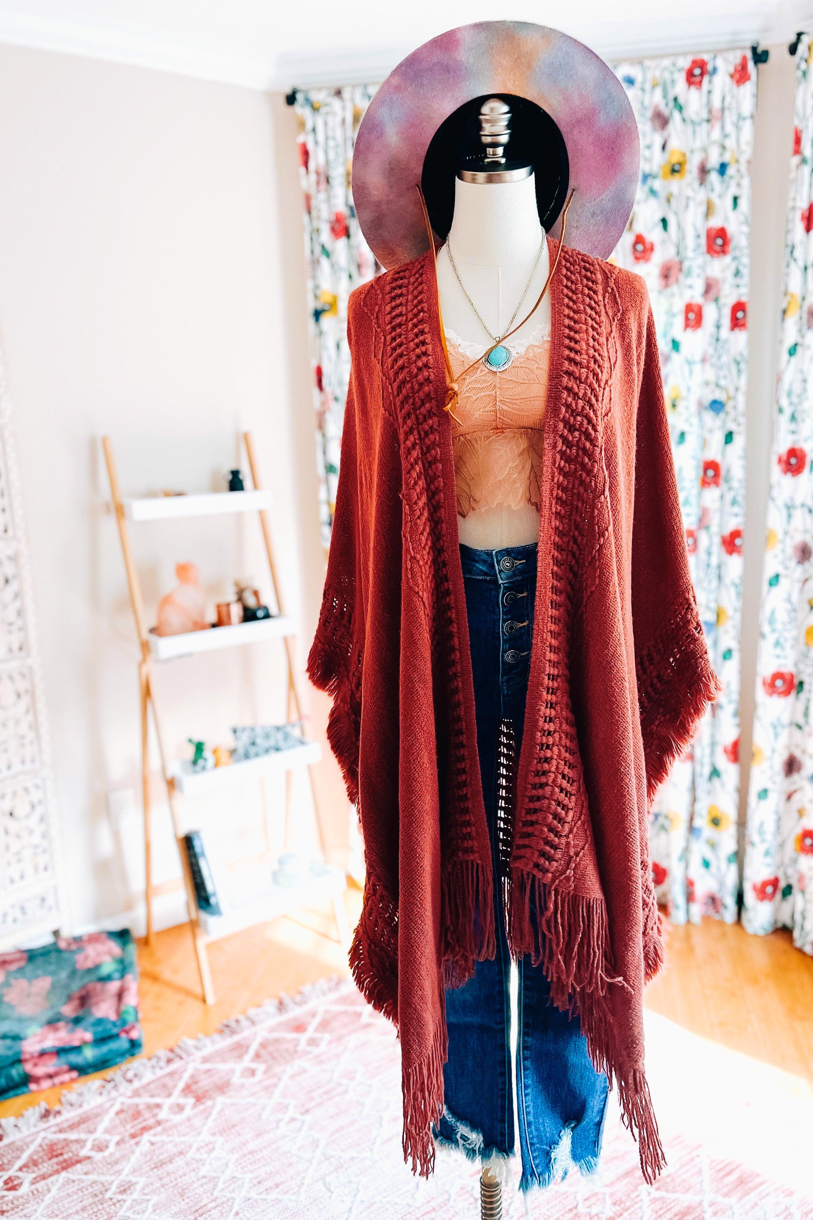 Mannequin dressed in rust-colored bohemian ruana with fringe, lace top, layered necklaces, and wide-brim hat indoors