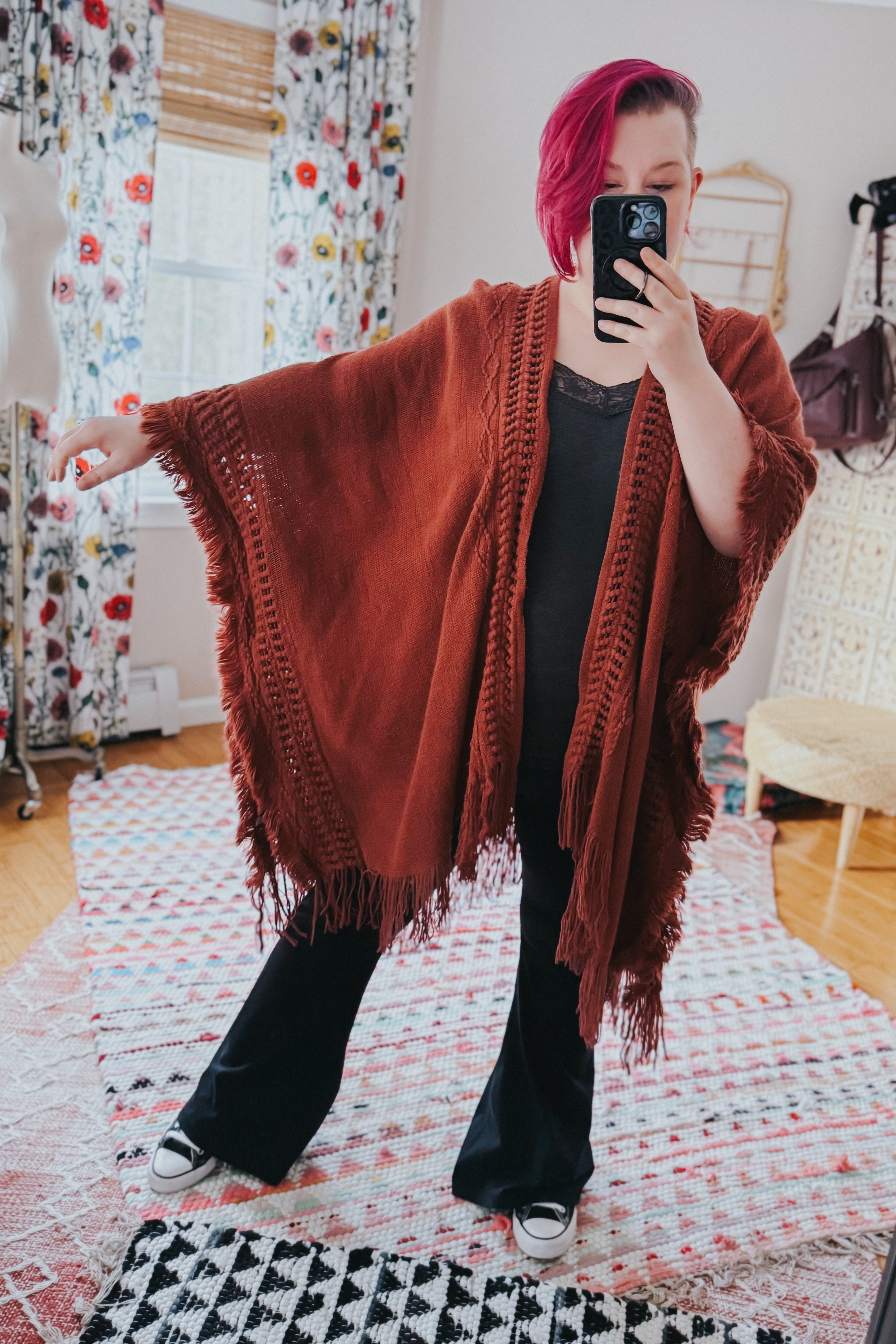 Woman wearing a rust-colored bohemian ruana with frayed edges over black outfit in a bright eclectic room