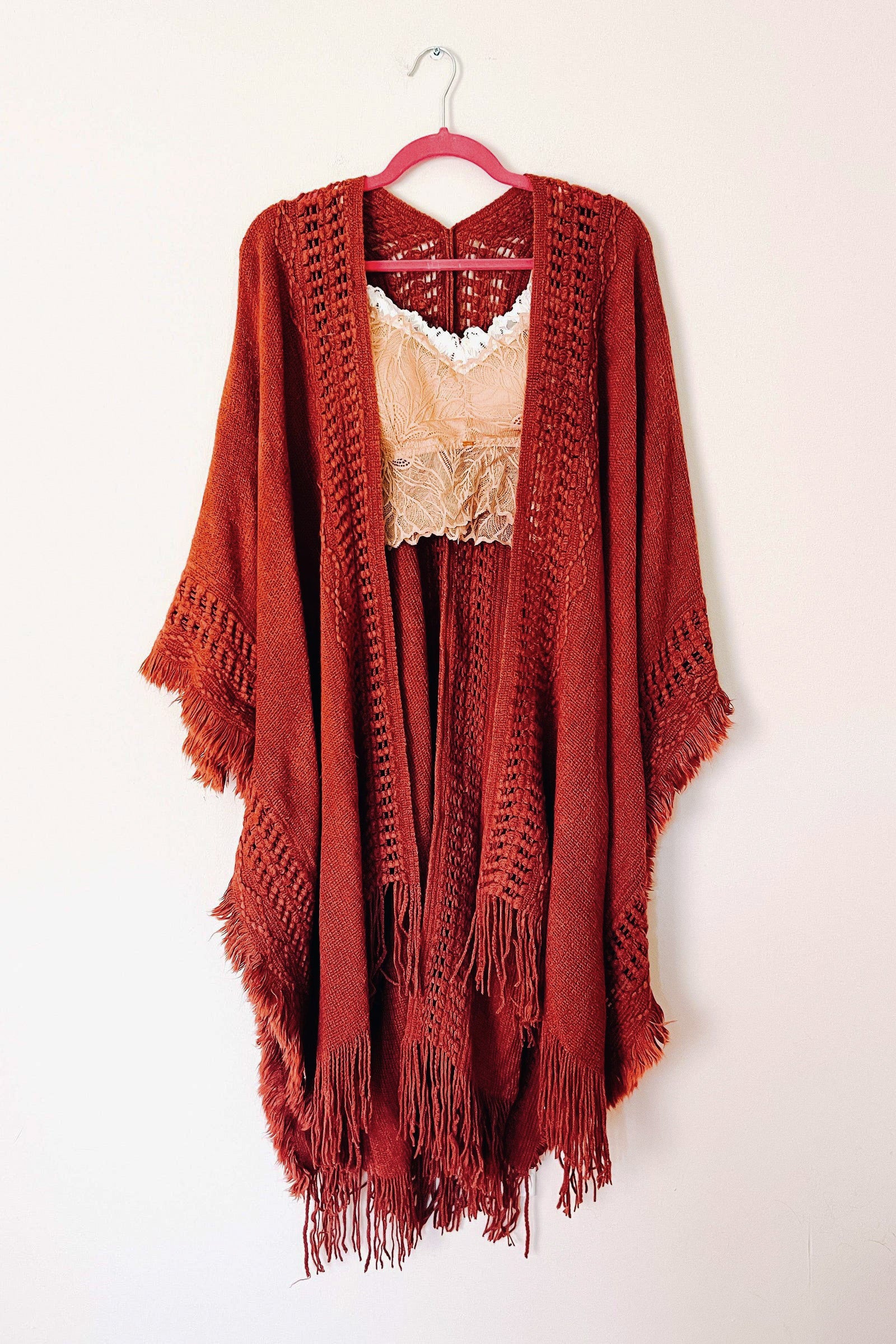 Rust-colored bohemian ruana with frayed edges and woven details layered over a beige lace top on a hanger