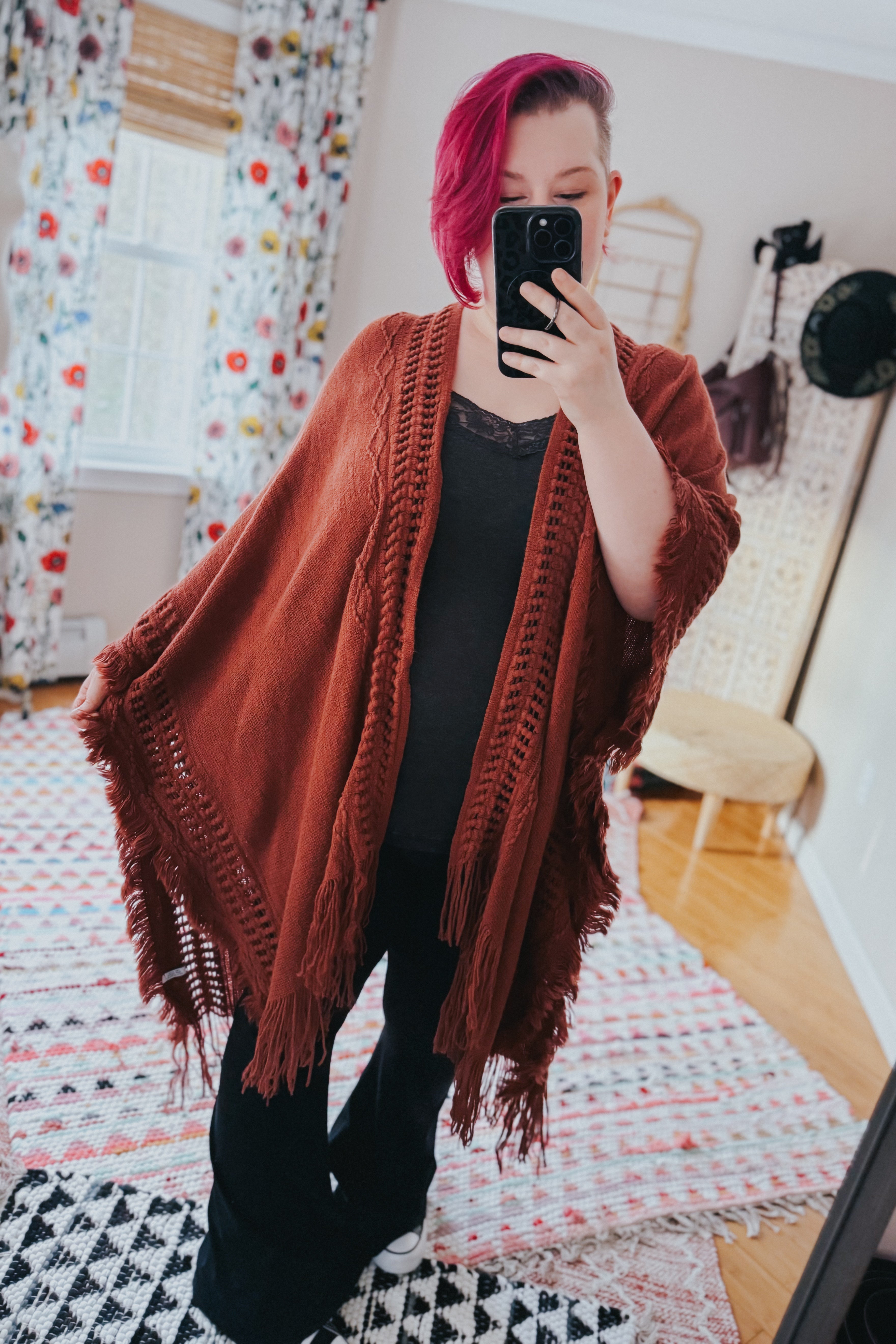 Woman wearing a rust-colored bohemian ruana with frayed edges over black outfit in a bright eclectic room