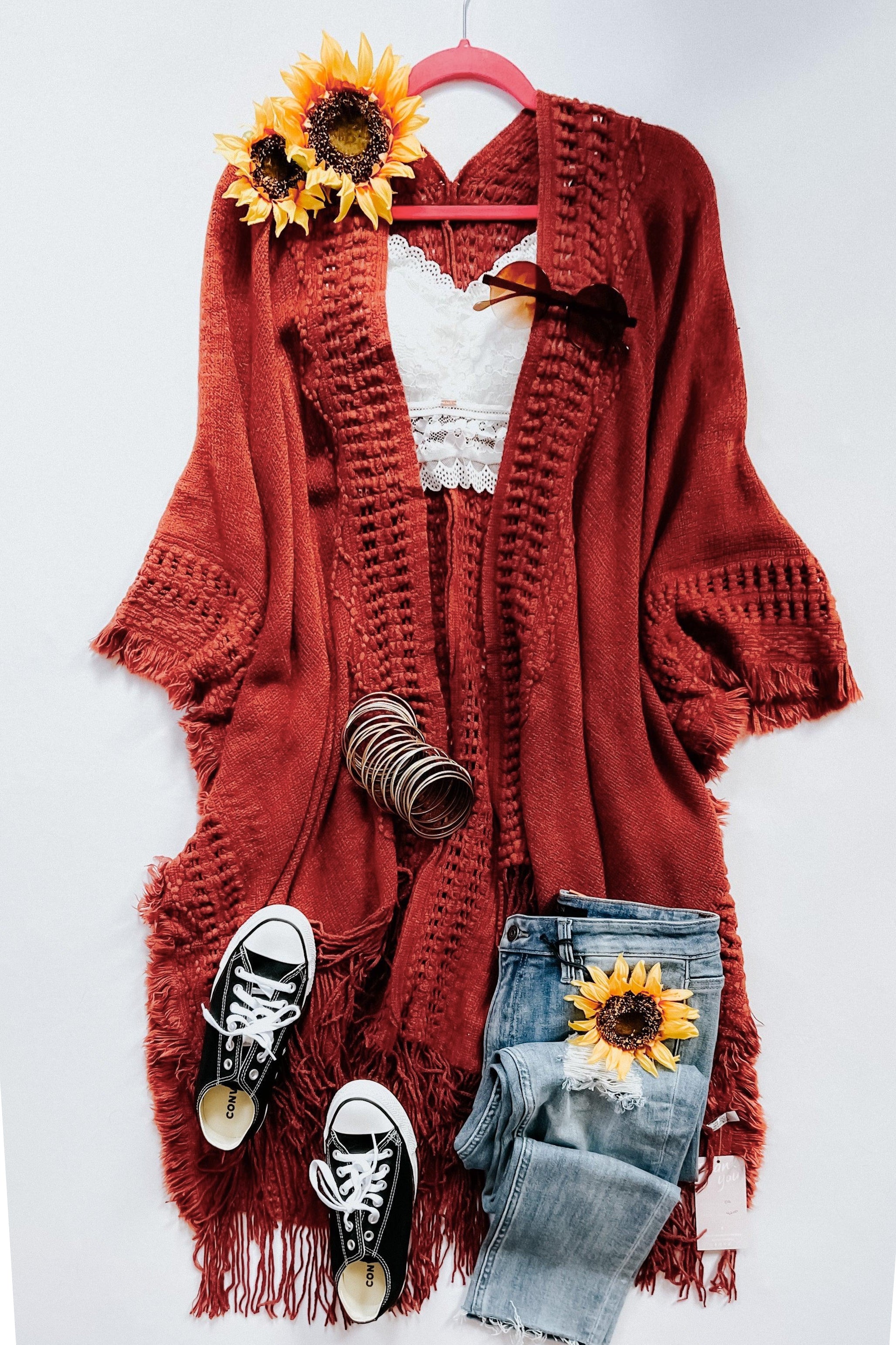 Rust-colored bohemian ruana with fringe paired with lace top, distressed jeans, black sneakers, and sunflower accents