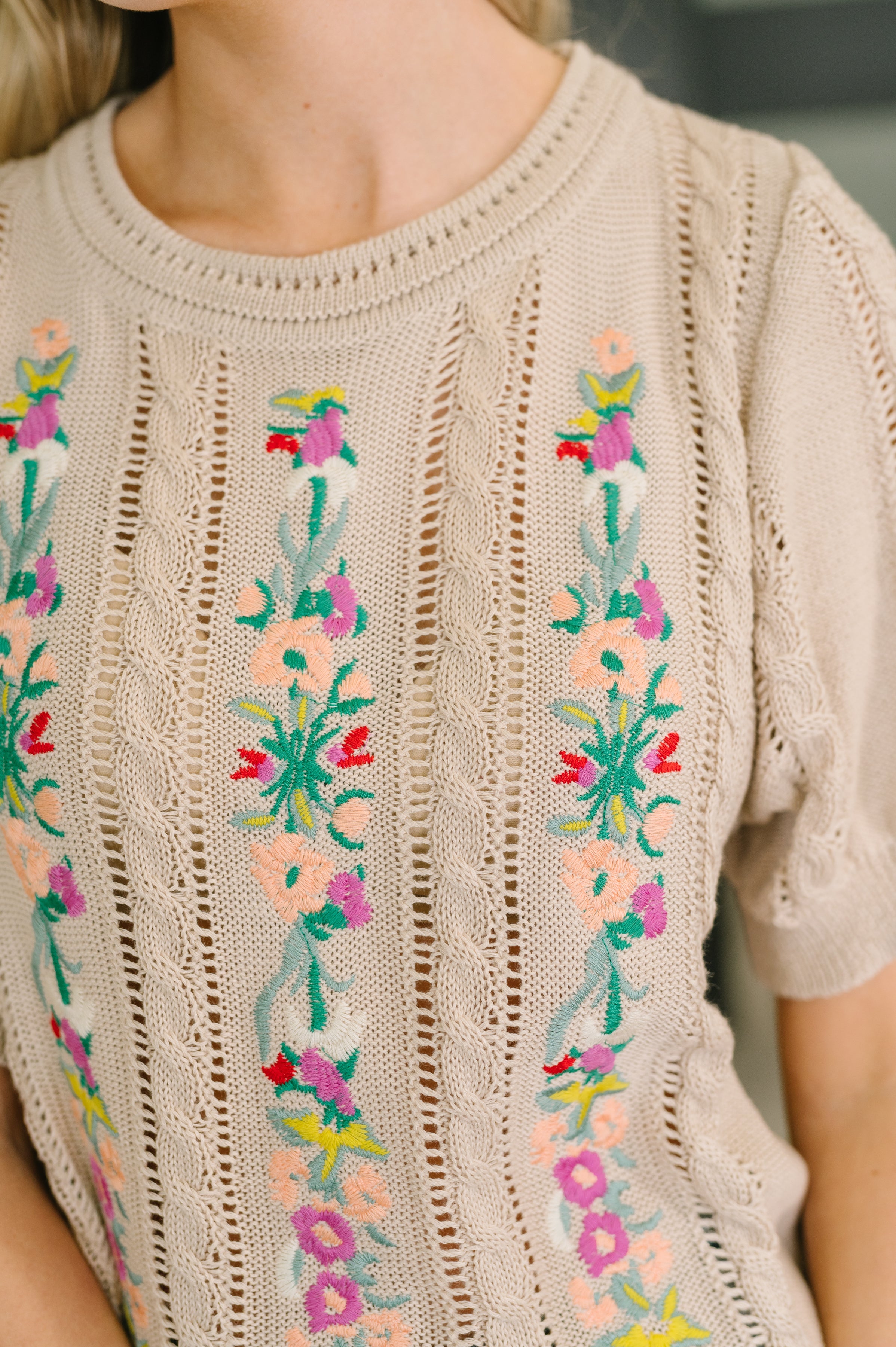 Close-up of beige knit top with intricate floral embroidery and open-knit vertical patterns in natural light