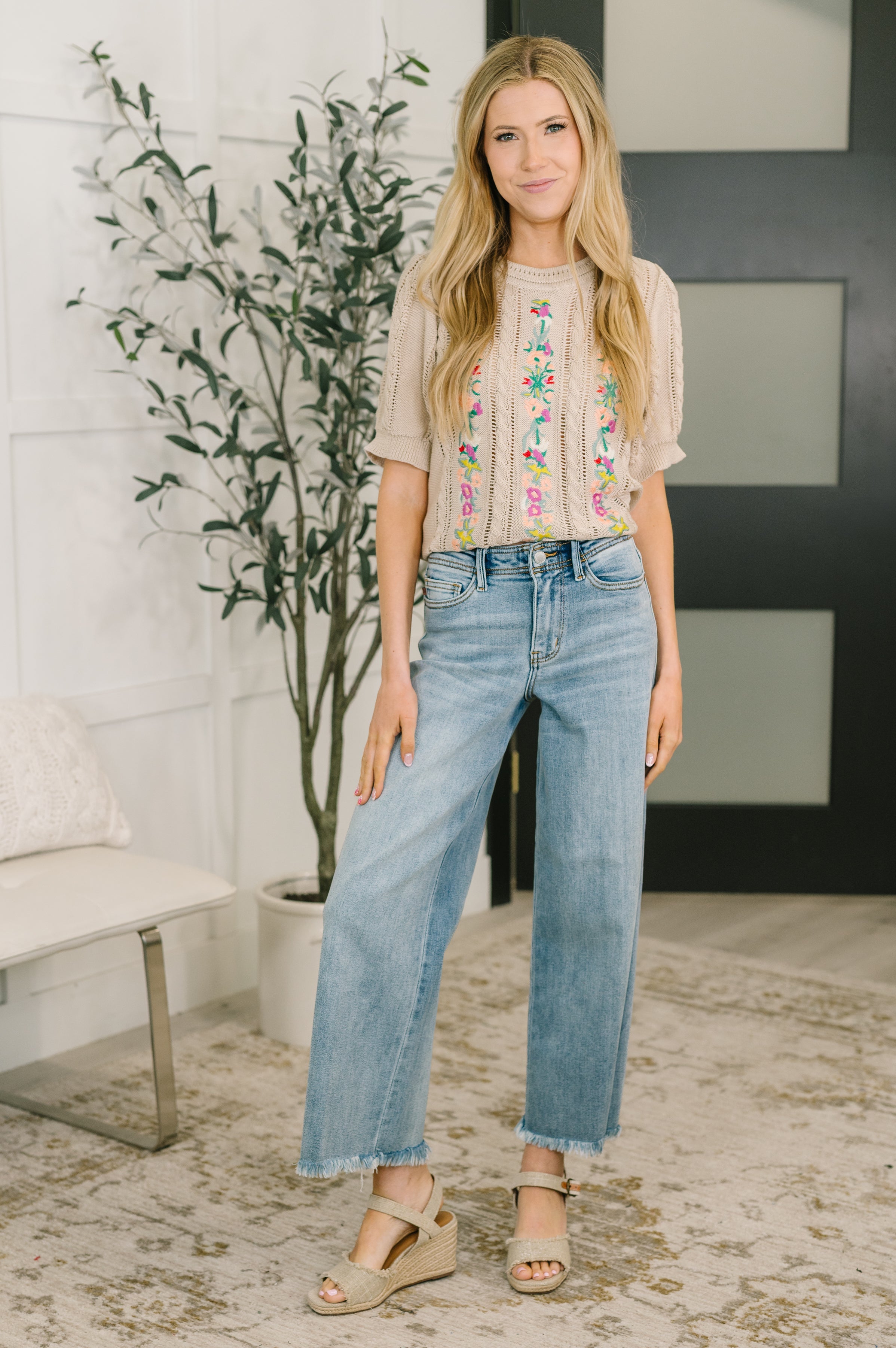 Woman wearing beige knit top with colorful embroidery paired with light wash straight jeans and wedge sandals indoors