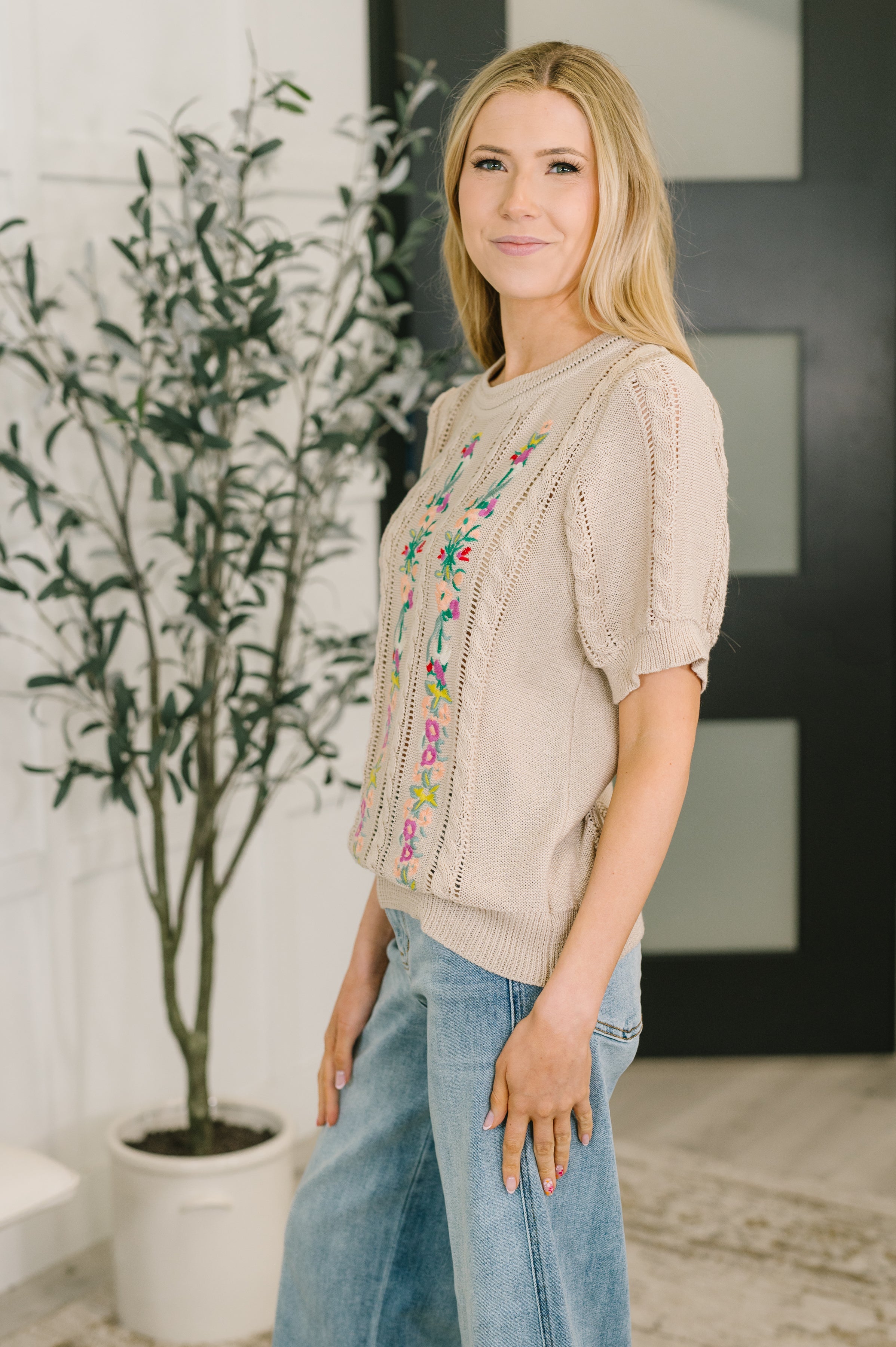 Woman wearing beige embroidered knit top with puff sleeves paired with light blue wide-leg jeans indoors