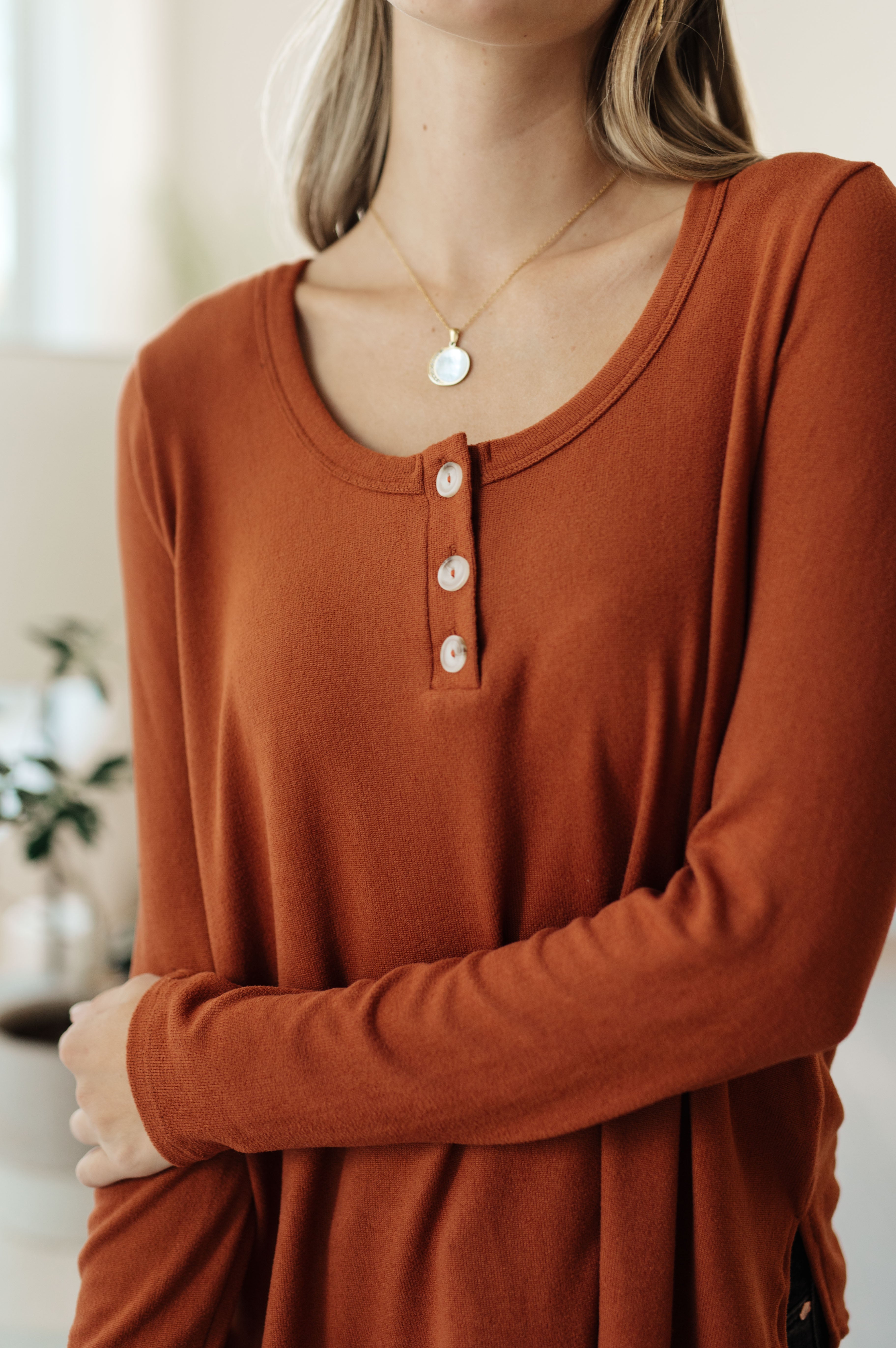 Close-up of a woman wearing a rust scoop neck long sleeve top with three-button placket and delicate gold pendant necklace