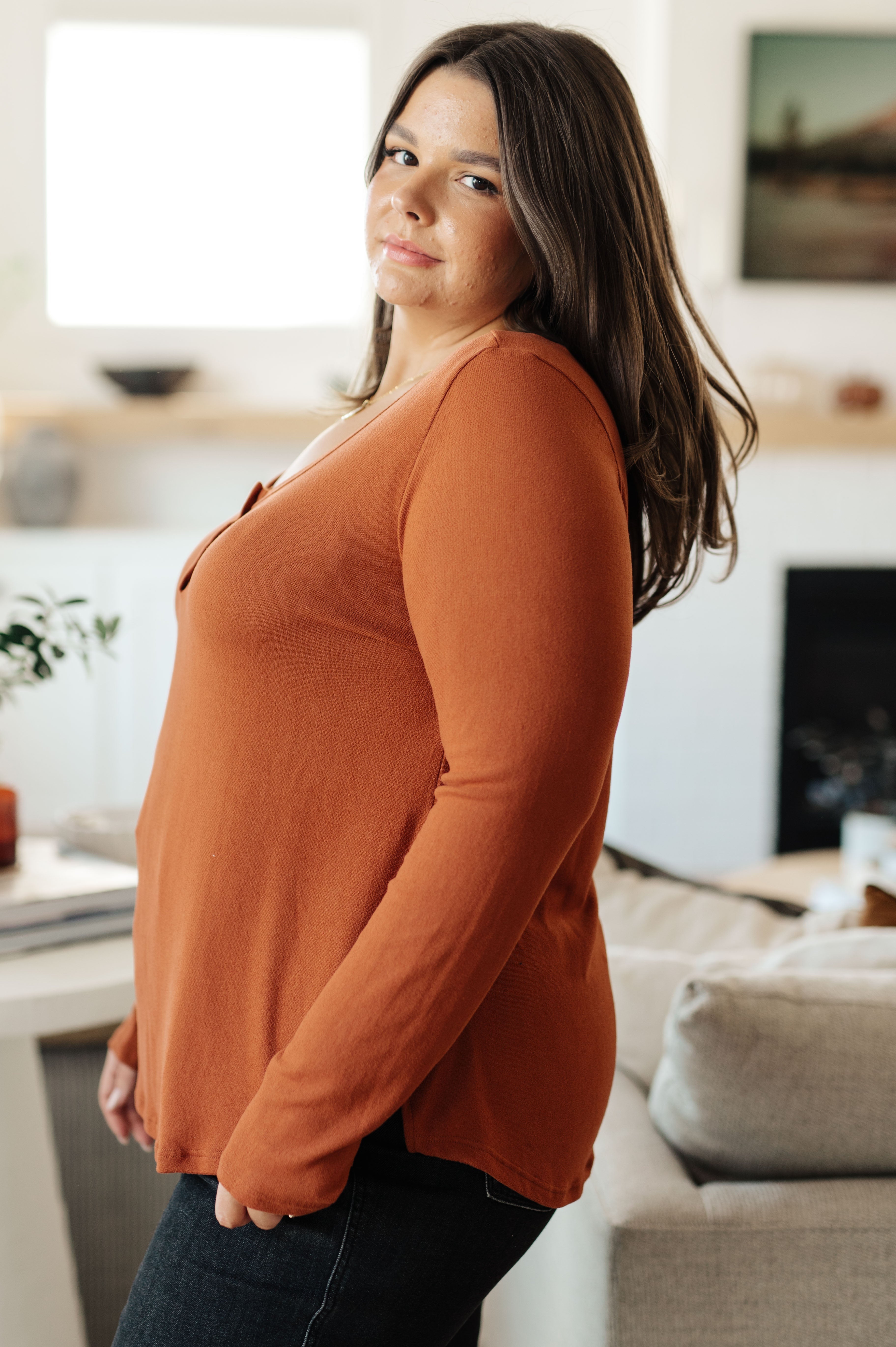 Side profile of woman wearing a rust scoop neck long sleeve top in a bright, home interior setting