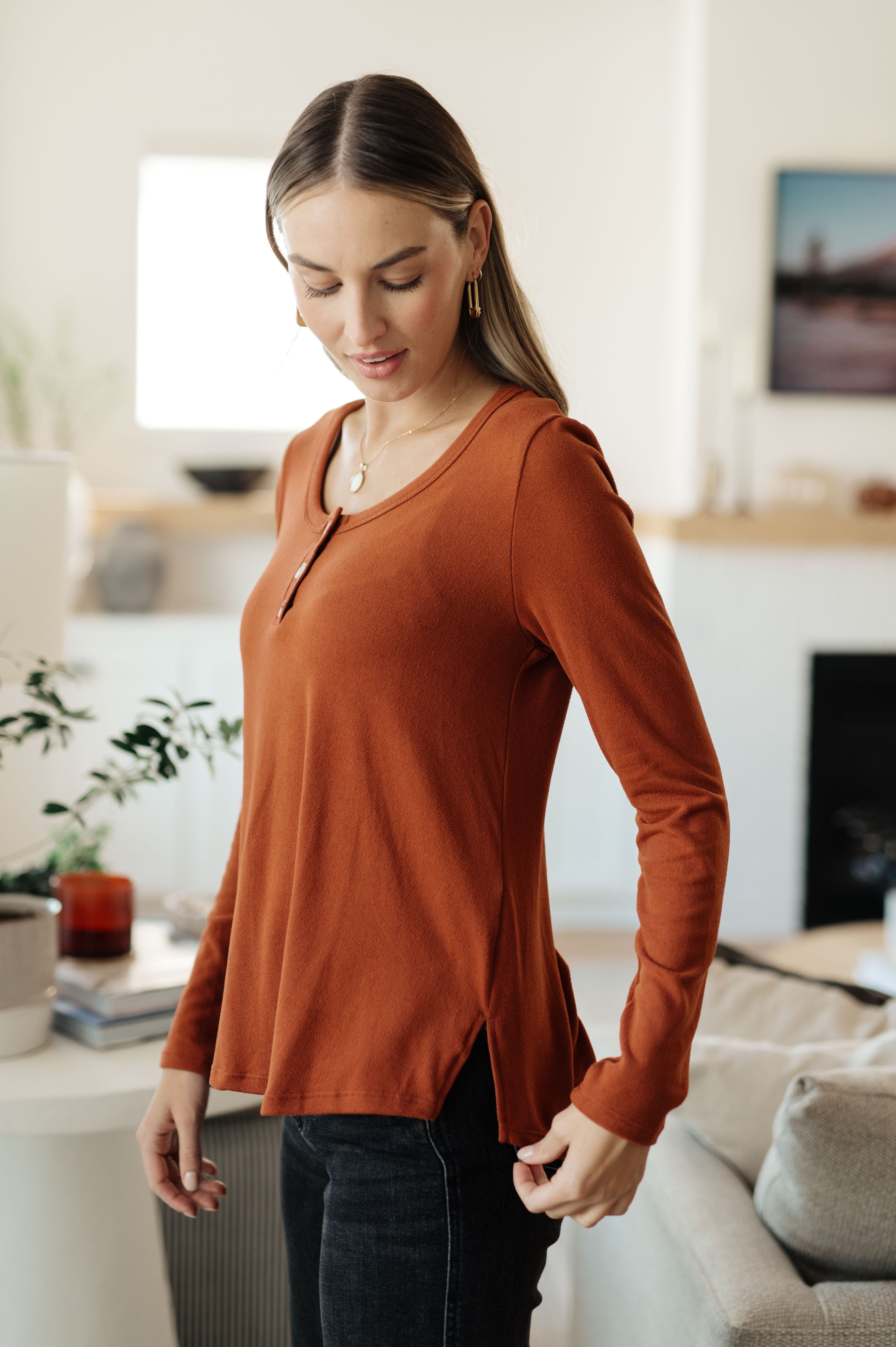 Woman wearing a rust scoop neck top with side slits and button details in a bright cozy living room setting