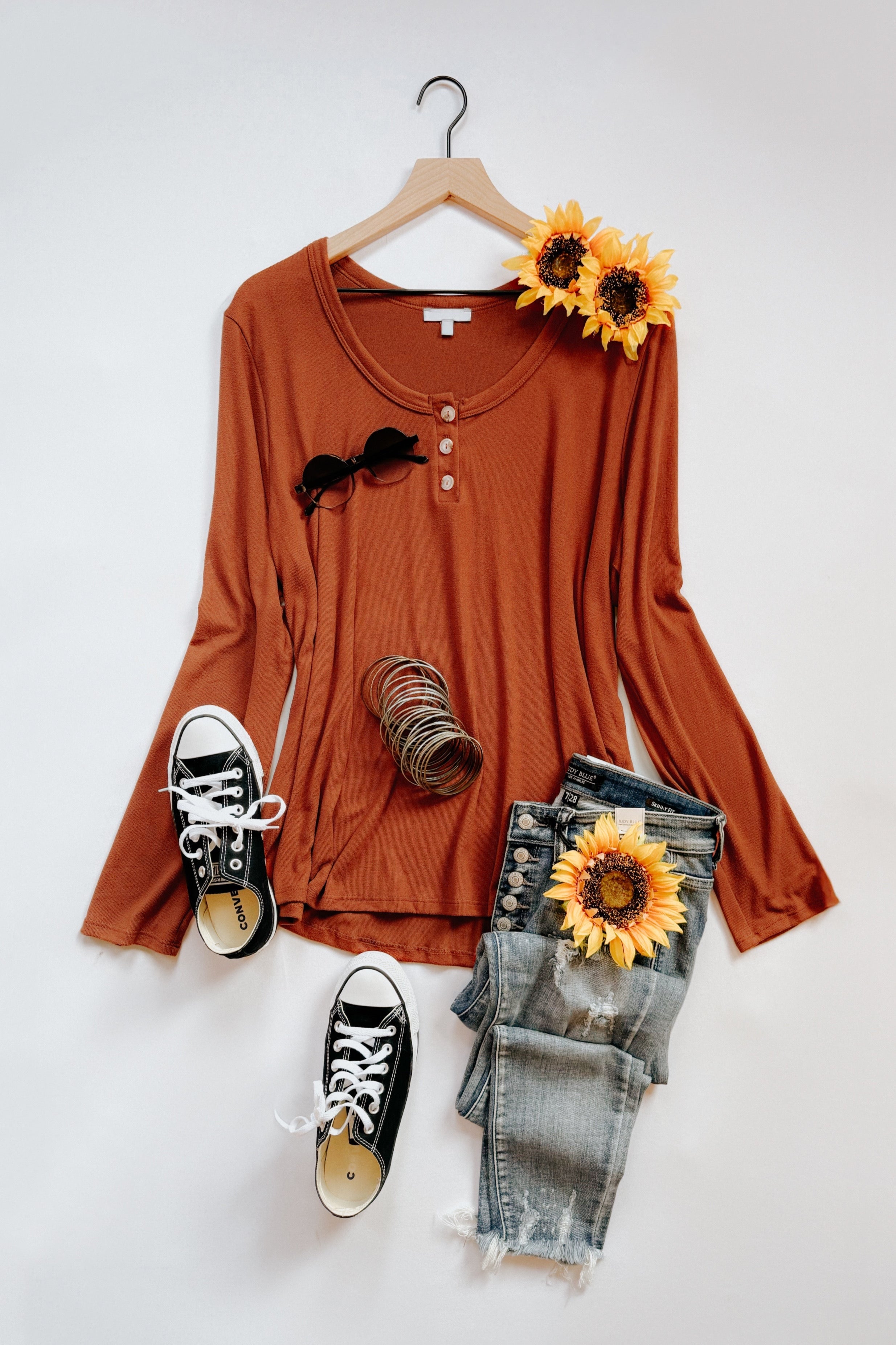 Rust scoop neck long sleeve top with button details styled with black sneakers, distressed jeans, and sunflowers flat lay