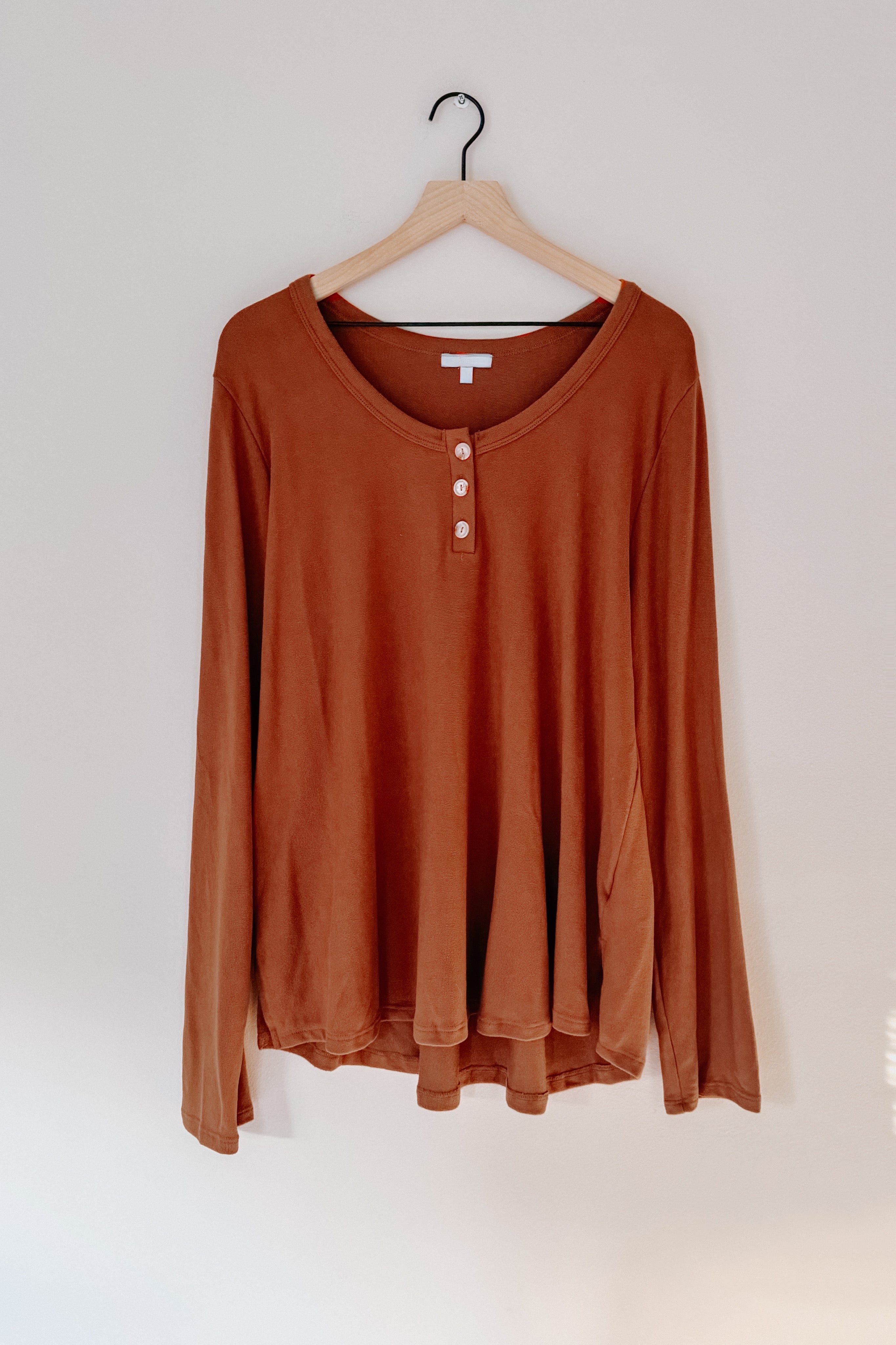 Rust-colored long sleeve scoop neck top with three button accents hanging on wooden hanger against white wall