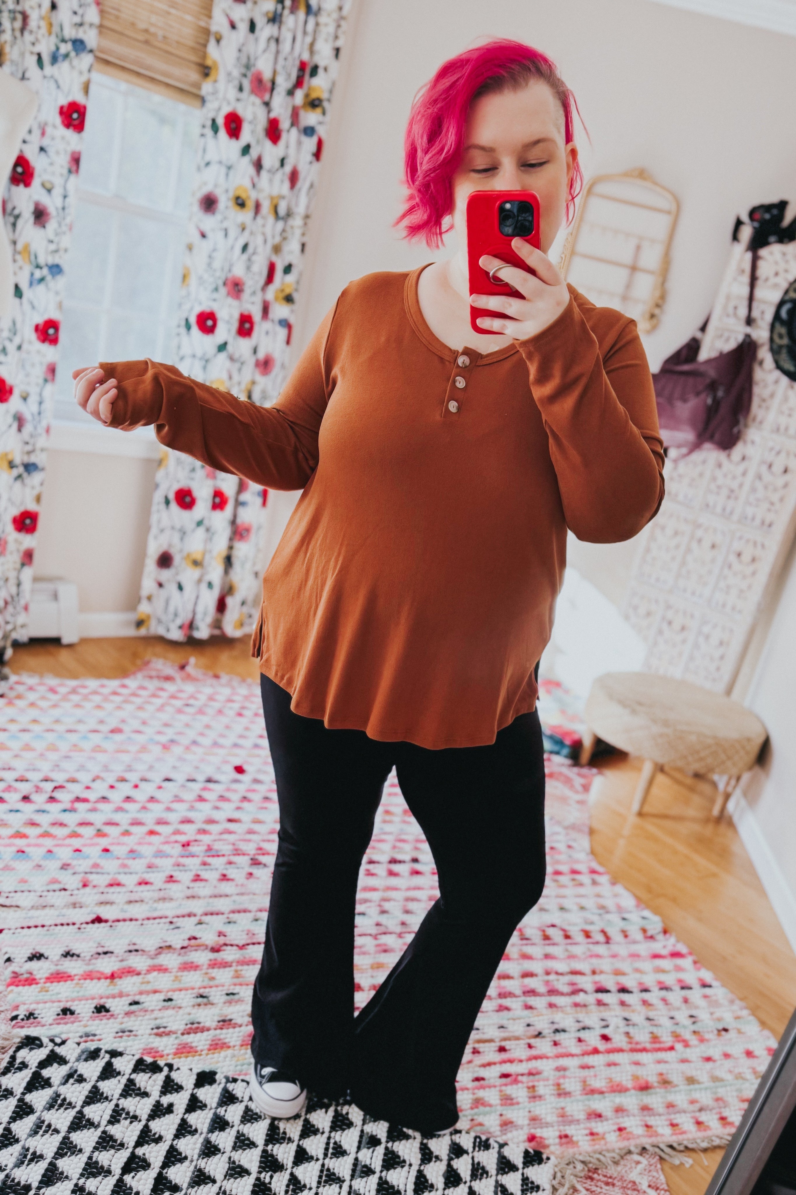 Woman with pink hair wearing a rust scoop neck top with buttons and black pants taking a mirror selfie in a bright room