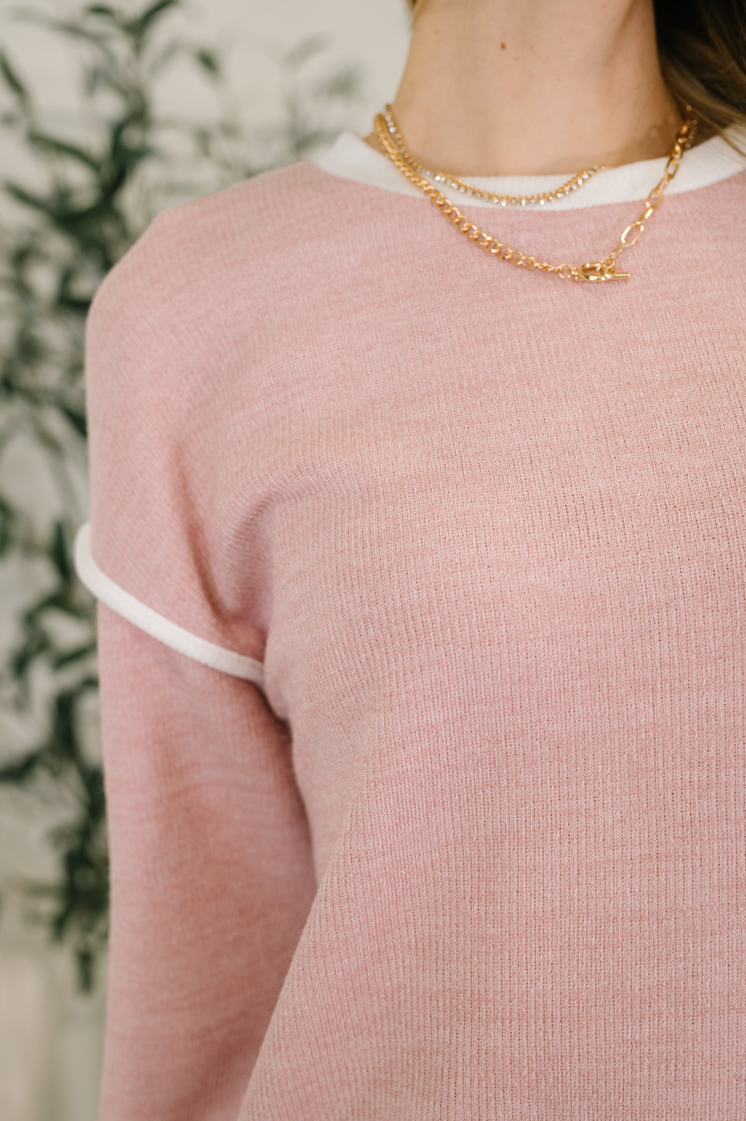 Close-up of woman wearing soft pink textured drop shoulder top with white trim and gold chain necklaces indoors