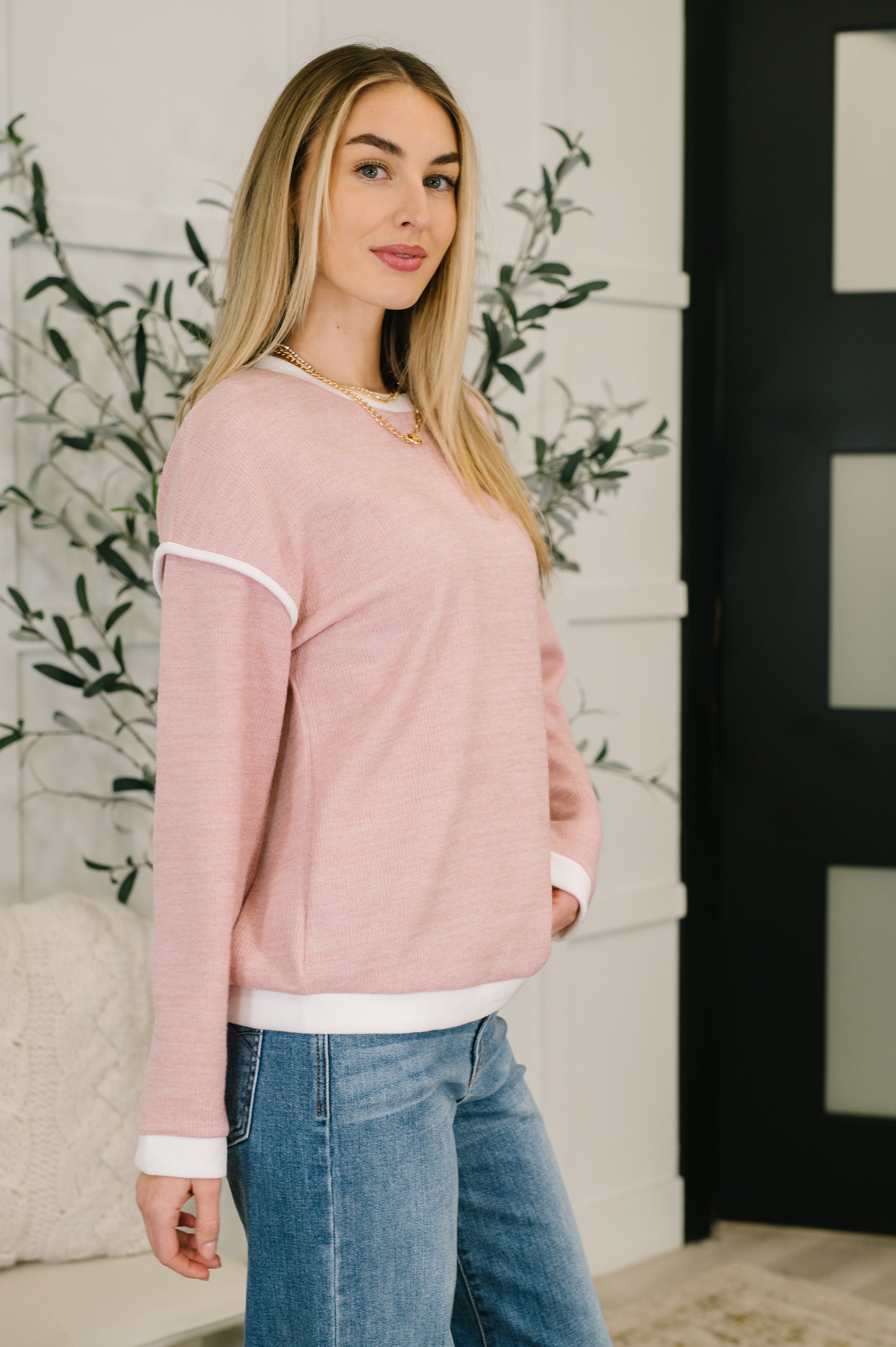 Woman wearing a light pink textured drop shoulder top with white trim and blue jeans indoors